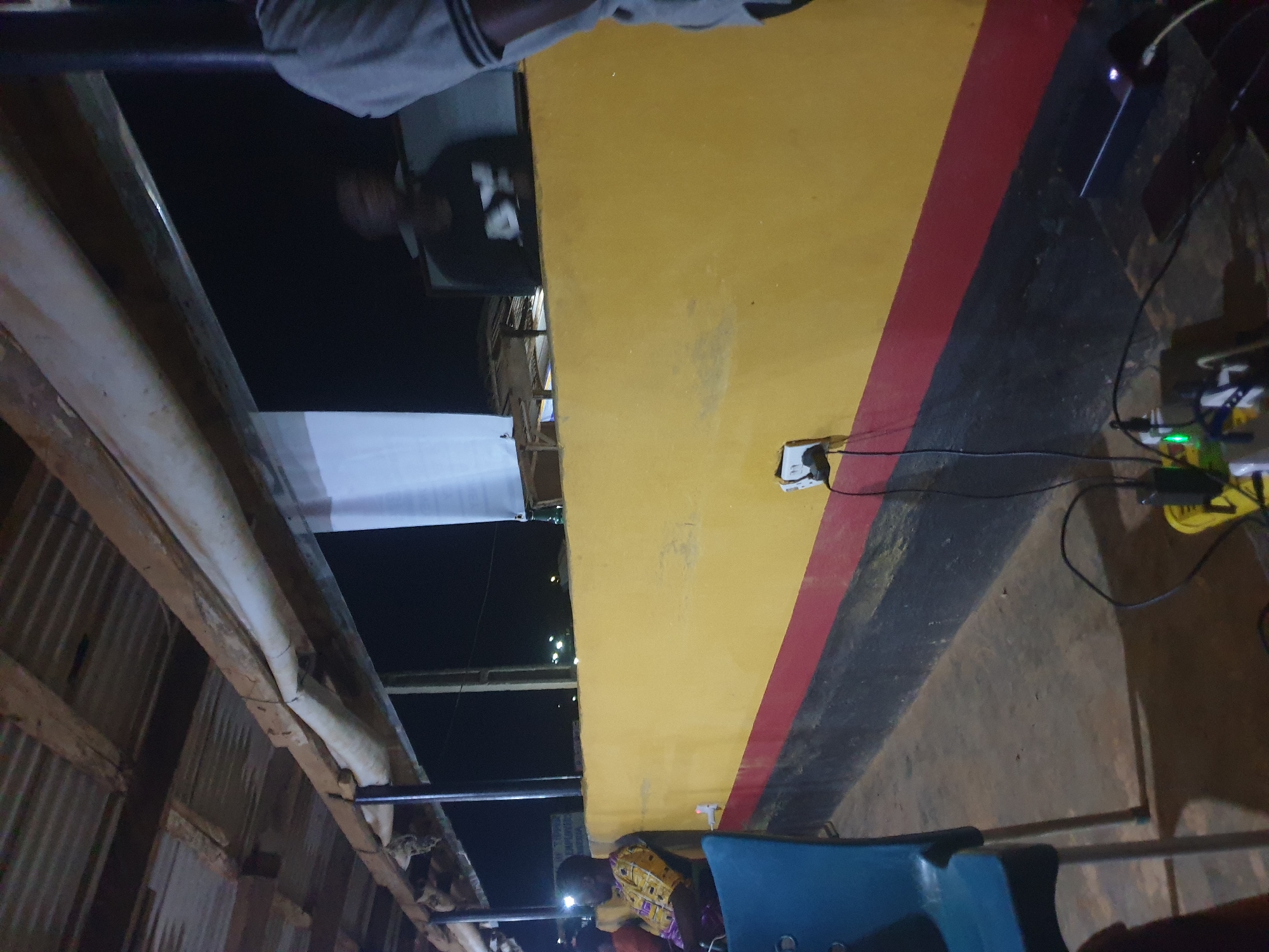 The image depicts an outdoor scene at night in Anilelerin, Nigeria. In the foreground, a blue chair is partially visible. Behind it, a person wearing a patterned shirt is seated, looking away from the camera. The middle ground features a yellow wall with black and red stripes along its base. An electrical outlet is mounted on the yellow wall, with wires connecting to various devices on the ground, including a power strip with illuminated green lights and a black rectangular device. In the background, the silhouettes of people are visible against a dark, possibly outdoor setting. Above the yellow wall, a corrugated metal roof with wooden beams is supported by pillars. The overall environment appears to be an open-air gathering space or shelter.