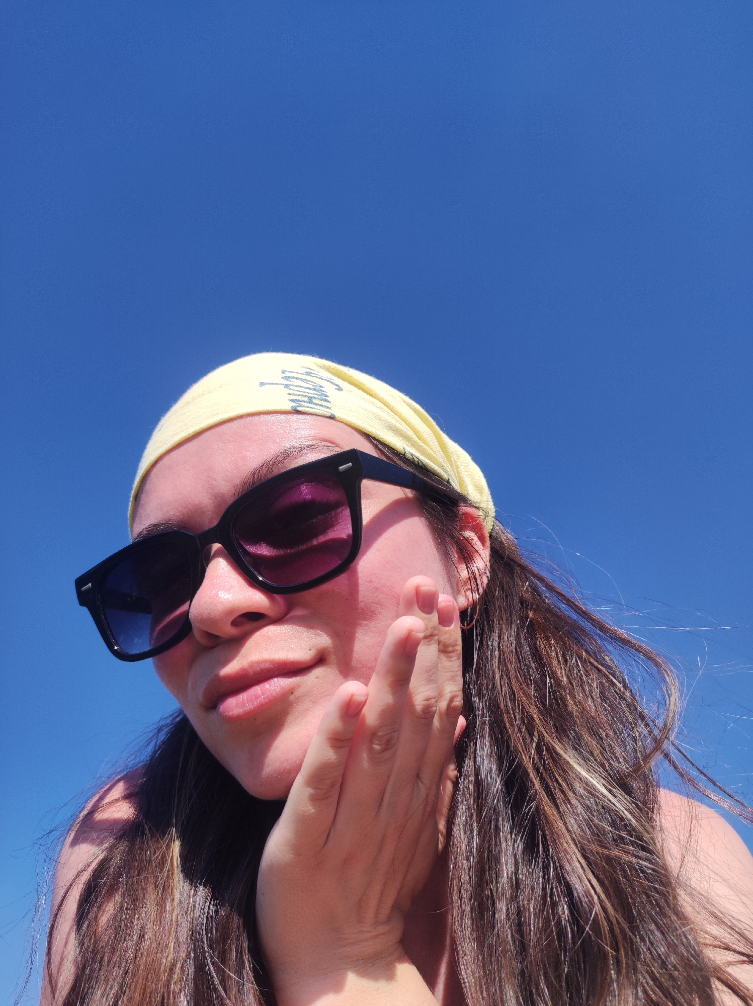 A selfie-style, low-angle shot shows a person with brown hair and fair skin, wearing sunglasses and a yellow headscarf against a bright blue sky. The person's left hand is touching their cheek. No specific details within the image confirm a location in Фоки, Russia.
