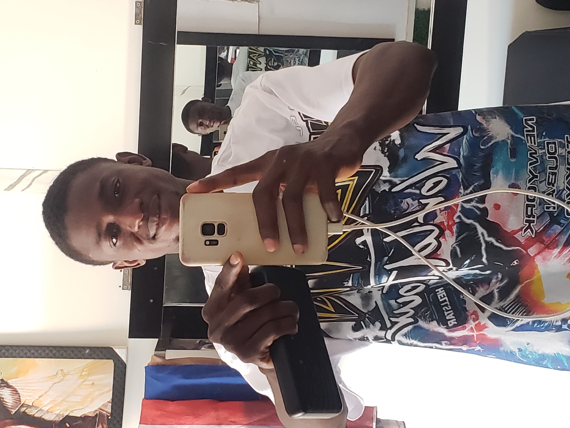 A young man is depicted taking a selfie in an indoor setting in Adankolo, Nigeria. He holds a light-colored smartphone in his right hand, pointed towards himself, and a dark, rectangular object (possibly a power bank) in his left. His reflection is prominent in a large, dark-framed mirror positioned behind him.
The man wears a white t-shirt adorned with a vibrant, multi-colored graphic design. Visible text on his shirt, reversed due to the mirror, includes "NEW YORK" and "ONEOFTHEBEST," along with other reversed characters such as "NOM" and "HEITZVK" amidst abstract patterns. A white charging cable is connected to his phone.
The background consists of a plain white wall, a framed artwork with warm, earthy tones (yellow, orange, brown), and a stack of items in blue, white, and red, which could be fabric or a flag. The man has short dark hair and a neutral to slightly smiling expression as he looks towards the camera. The indoor lighting suggests a daytime setting.