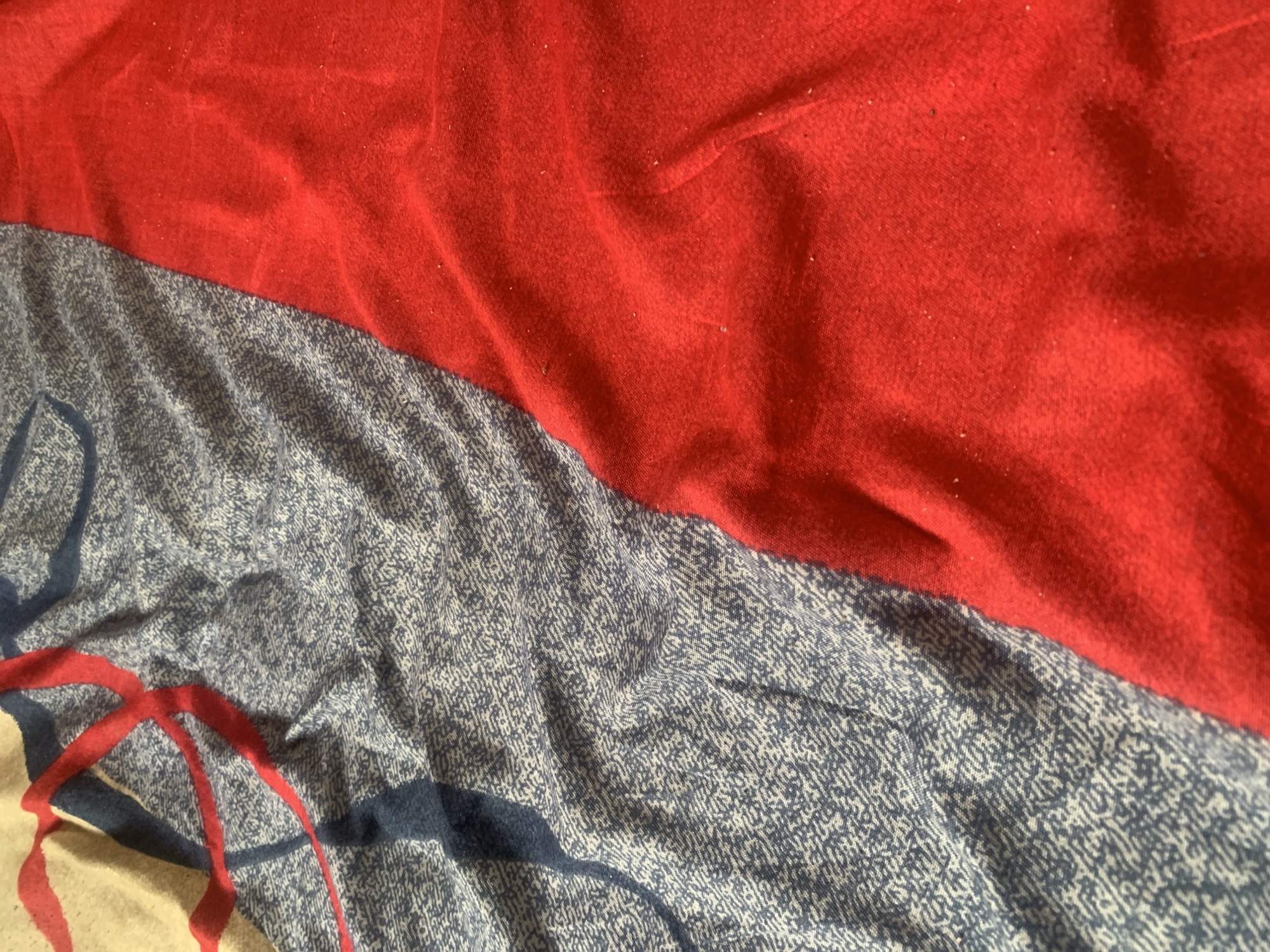 The image is a close-up, overhead shot of a crumpled fabric, likely a blanket or bedsheet, divided into two main sections by a diagonal seam. The left side features a patterned fabric with a light grey background and a dense, repeating dark blue print that resembles small, irregular shapes. Overlaying this pattern are thick, somewhat fuzzy red lines and thinner, solid dark blue lines that form abstract shapes, possibly part of a larger design. The right side of the fabric is a solid, vibrant red and appears to be a different material, possibly softer and more textured, with visible lint or small fibers scattered across its surface. The lighting suggests it is an indoor setting, with shadows creating depth and highlighting the folds and textures of the fabric. The image does not depict any people, actions, or interactions. There is no discernible connection to the city of Jauro Yinu, Nigeria, as the image focuses solely on the fabric's texture and pattern.