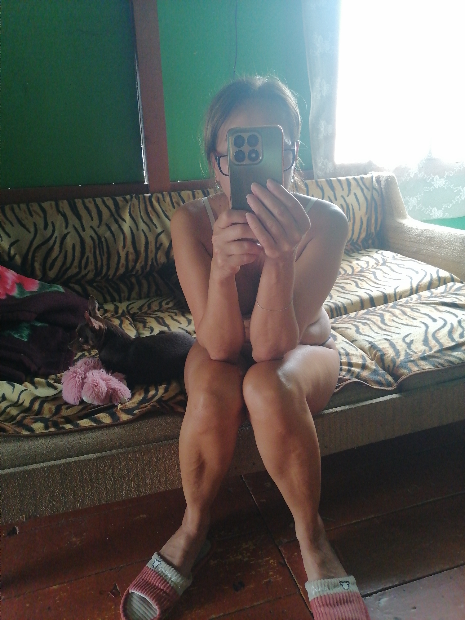 A woman is taking a mirror selfie in a room. She is sitting on a sofa with a tiger print couch cover, holding a smartphone in front of her face. She is wearing glasses and a white top. A small dog is lying on the sofa next to her, near a pink fluffy toy. The room has green walls and wooden floors. Natural light is coming in from a window on the right side of the frame.