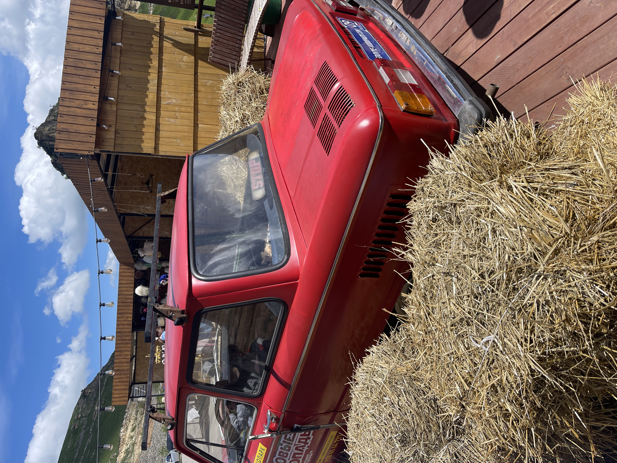 The image shows a red vintage car, possibly a Zaporozhets, nestled amongst bales of hay. The setting appears to be outdoors in Красный Курган, Russia, with a partly cloudy sky visible in the background along with mountains. A wooden building and decorative string lights are also present. The car has a roof rack and signage in Russian. No people are clearly visible, although there might be individuals reflected in the car's windows.