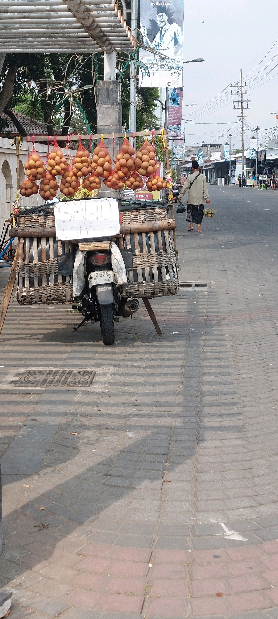The image depicts a street scene with a motorcycle parked on a paved walkway. The motorcycle is loaded with a large woven basket structure, which extends significantly from its sides. Hanging from a wooden frame above the basket are several netted bags filled with what appear to be fruits, likely sawo given the sign on the basket. The sign, written in white on a dark background, reads "SAWO MANIS."
In the background, a person wearing a light-colored long-sleeved shirt, a dark sarong, and a black cap walks away from the camera, carrying a dark bag in their left hand and a small basket with some items in their right hand. Further down the street, more people are visible, as are several buildings lining the road, suggesting a commercial or market area. Overhead, power lines and poles crisscross the sky, and a large poster featuring a portrait of Ir. Soekarno is prominently displayed on a pole. The overall environment suggests a street in a tropical or subtropical region, with the architecture and the presence of street vendors indicating a local marketplace. The pavement is made of interlocking bricks, and shadows of the fruit display and the motorcycle fall across it.