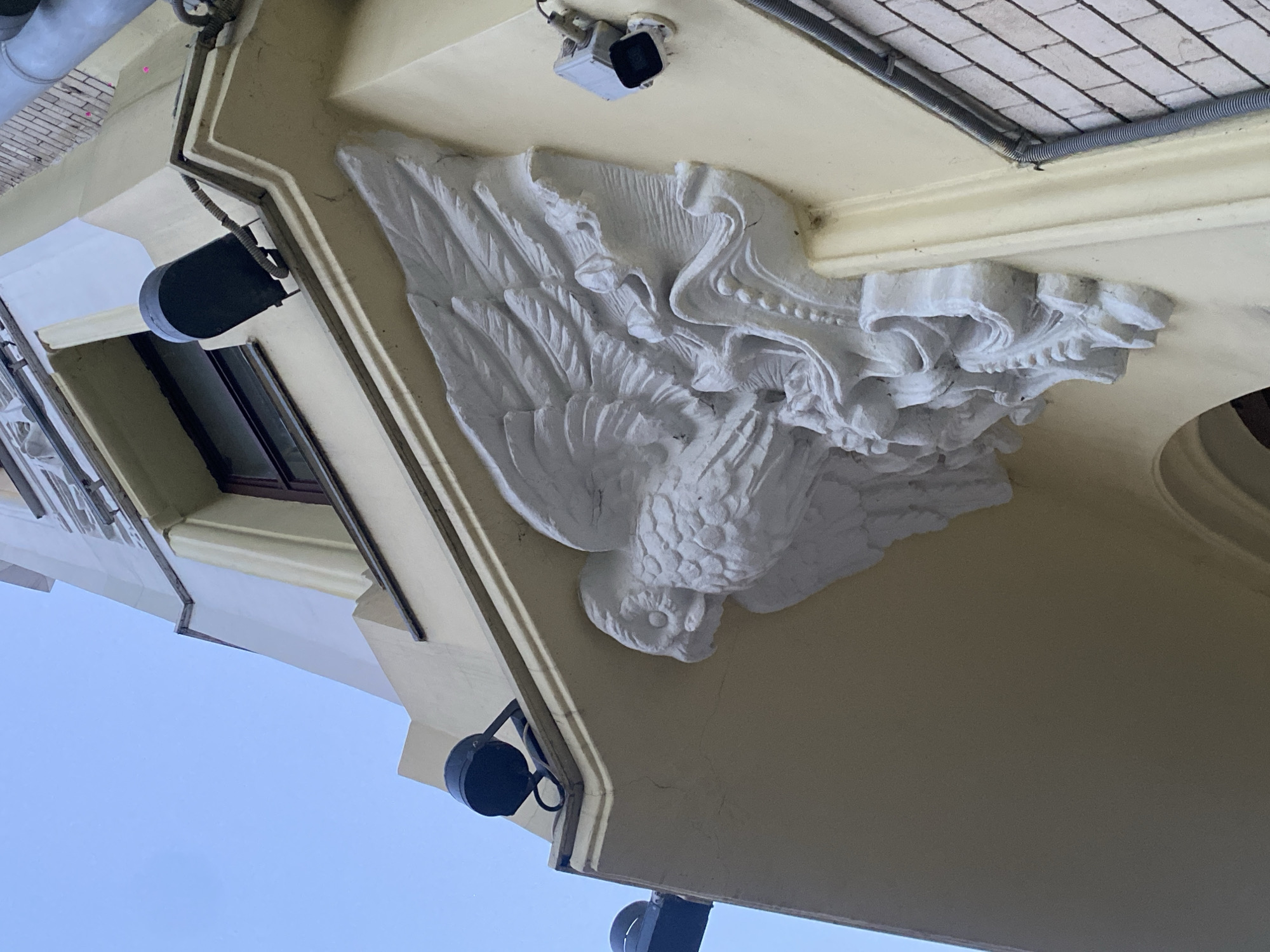 An upward-angled view captures the corner of the Art Nouveau-style Profitable House of Yakov Rechkin, located at Chistoprudny Boulevard, 21, building 1, Moscow, Russia. The building's facade is painted a light yellow. A prominent, white, high-relief architectural sculpture of an owl with outstretched wings is centrally positioned at the corner, resting above elaborate white scrollwork and foliage motifs. The owl's head is turned to the left. Above the sculpture, a dark-framed window is visible. Attached to the facade are several modern fixtures: two black cylindrical spotlights are mounted on the upper left and above the owl, respectively. A light-colored security camera with a dark lens is affixed to the right side of the corner, above the owl's right wing. A grey corrugated electrical conduit runs vertically along the far right edge of the frame, adjacent to a section of light-colored brickwork. The sky is clear blue.