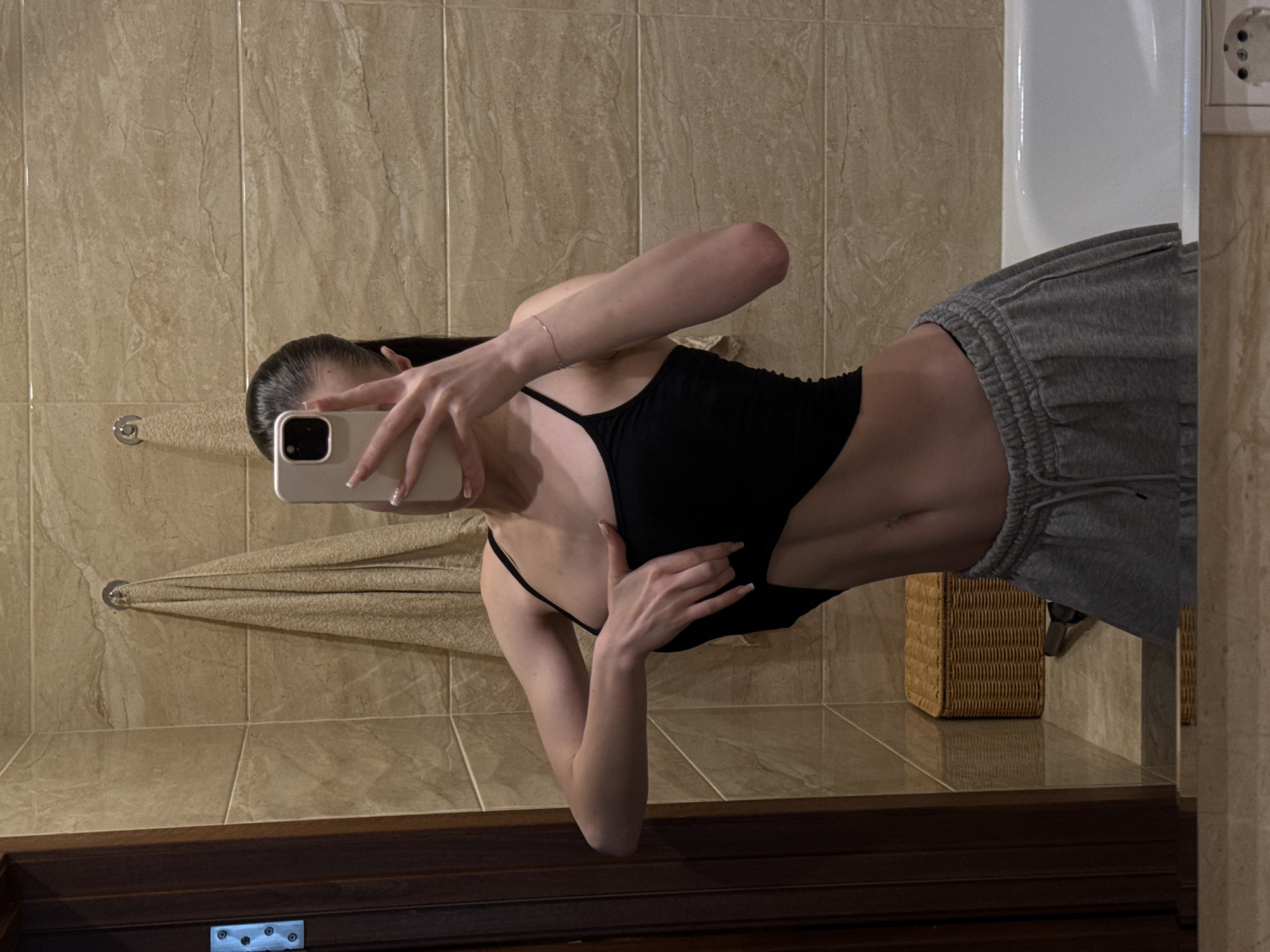 A young woman with dark hair is taking a mirror selfie in a bathroom. She is wearing a black crop top and grey sweatpants. Her left hand is resting on her stomach, and her right hand is holding a smartphone up to take the picture. The bathroom has tiled walls and a wicker basket on the floor. A white bathtub is visible in the background. The image is framed from the waist up. The photo was taken in the city of Khimki, Russia.