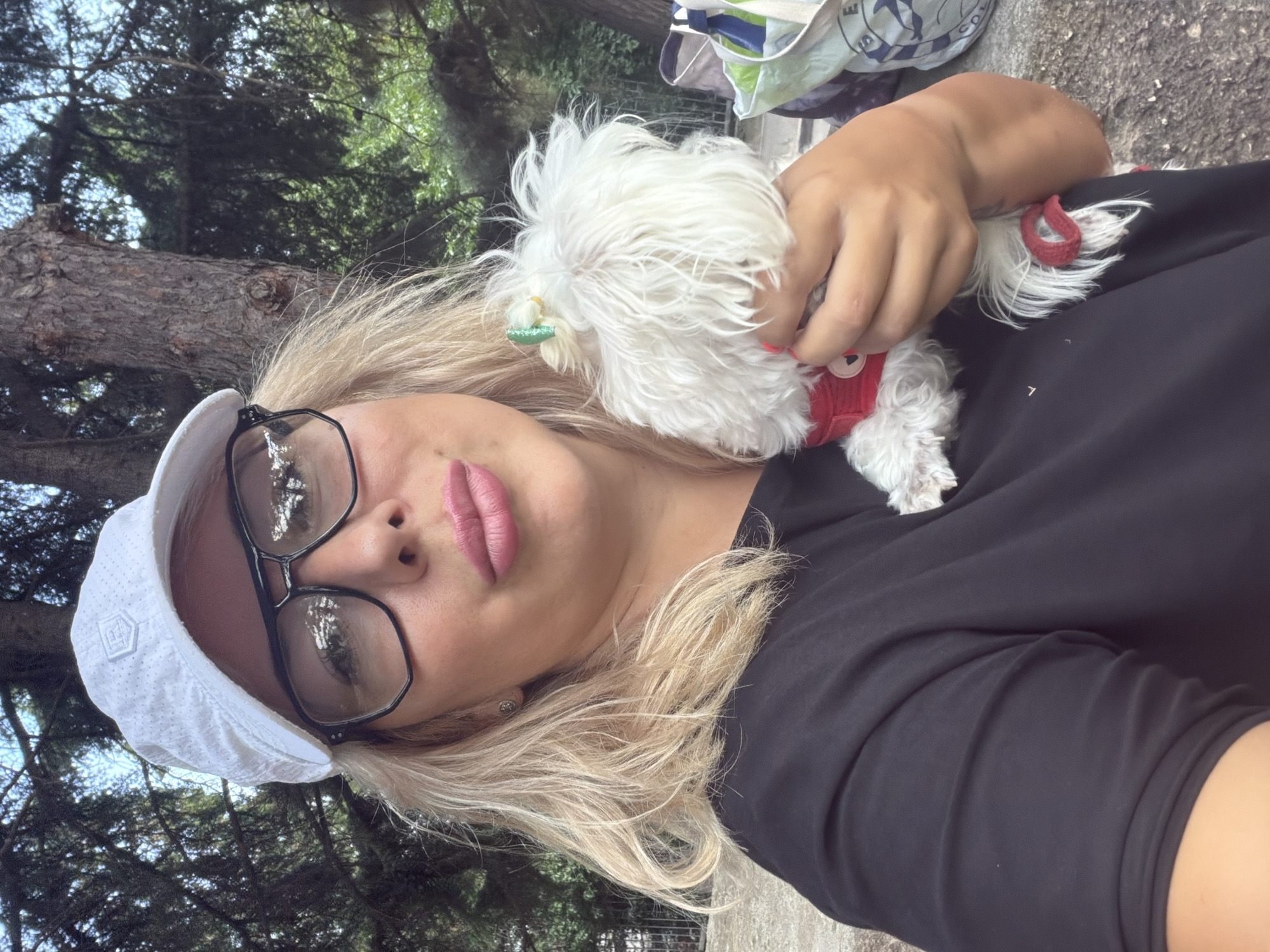 A blonde woman wearing a white baseball cap, black t-shirt, and large-framed glasses holds a small white dog. The dog is wearing a red harness with a small green bow on its head. They are outdoors, with trees and foliage in the background. The scene appears to be in a park or wooded area. The location is identified as Partenit, Ukraine.