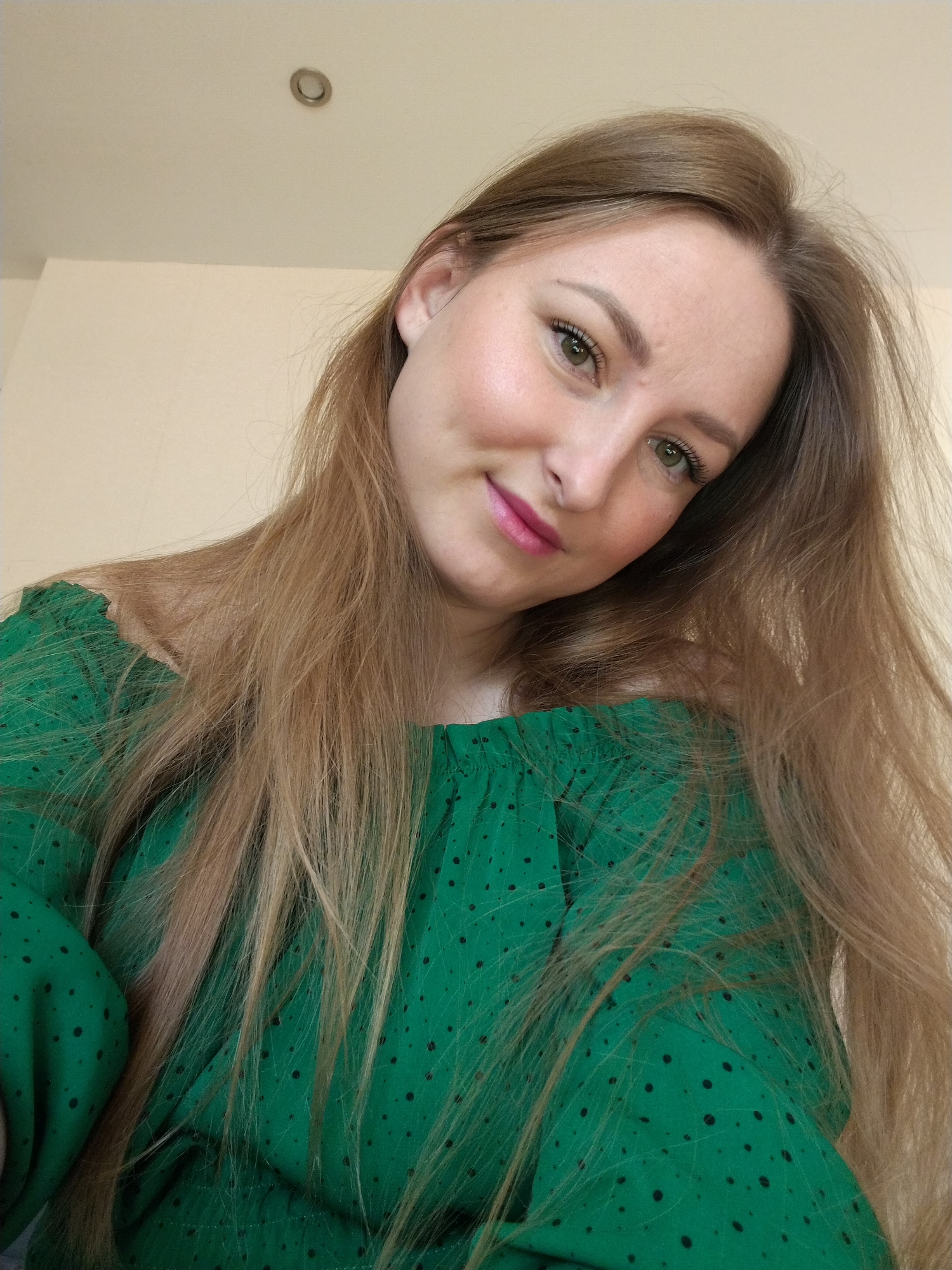 A young adult woman is featured in a close-up, head-and-shoulders portrait, likely a selfie. She has long, straight, light brown hair with blonde undertones that falls over her shoulders and chest. Her head is tilted slightly, and she looks directly at the camera with a gentle, friendly smile. She has fair skin, striking green eyes, and well-defined eyebrows. Her lips are adorned with bright pink lipstick, and a subtle blush is visible on her cheeks. She is wearing a vibrant green, off-the-shoulder top or dress, characterized by a pattern of small black polka dots and gathered fabric around the neckline.
The setting appears to be indoors, indicated by the plain, light beige or cream-colored walls and ceiling visible in the background. A single, round recessed light fixture is mounted on the ceiling in the upper-right portion of the frame. The lighting is soft and even, highlighting her features. The overall mood conveyed is calm and approachable. This image was captured in Chaikovsky, Russia.