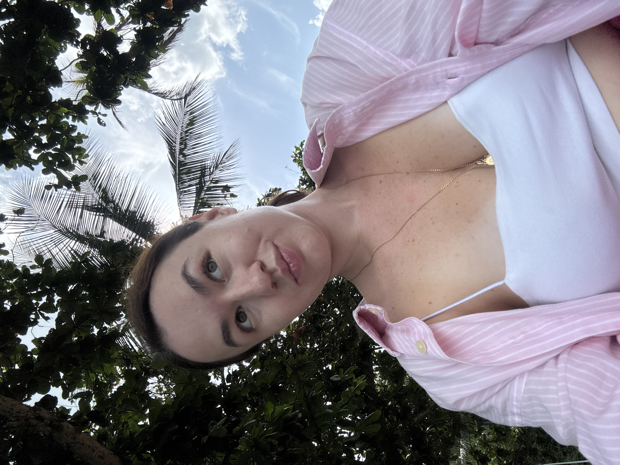 A woman is taking a selfie outdoors in Pa Khlok, Thailand. She is positioned in the lower center of the frame, looking up and to her right with a slight smile. She has fair skin with freckles and is wearing a white tank top and an open pink and white striped button-down shirt. A gold necklace with a pendant is visible. She has dark hair pulled back. The background consists of lush green foliage and palm fronds, with a bright blue sky and scattered white clouds visible through the leaves. The lighting suggests it is daytime, possibly late morning or early afternoon, with the sun filtering through the trees.