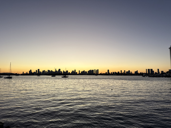 A cityscape silhouette stretches across the horizon, backlighting a harbor scene. Buildings of varying heights punctuate the sky, creating a distinct urban skyline. The water reflects the golden hues of the sunset, with boats scattered across the surface, enhancing the tranquil ambiance. The sky transitions from a warm orange to a cool violet, showcasing a serene sunset.