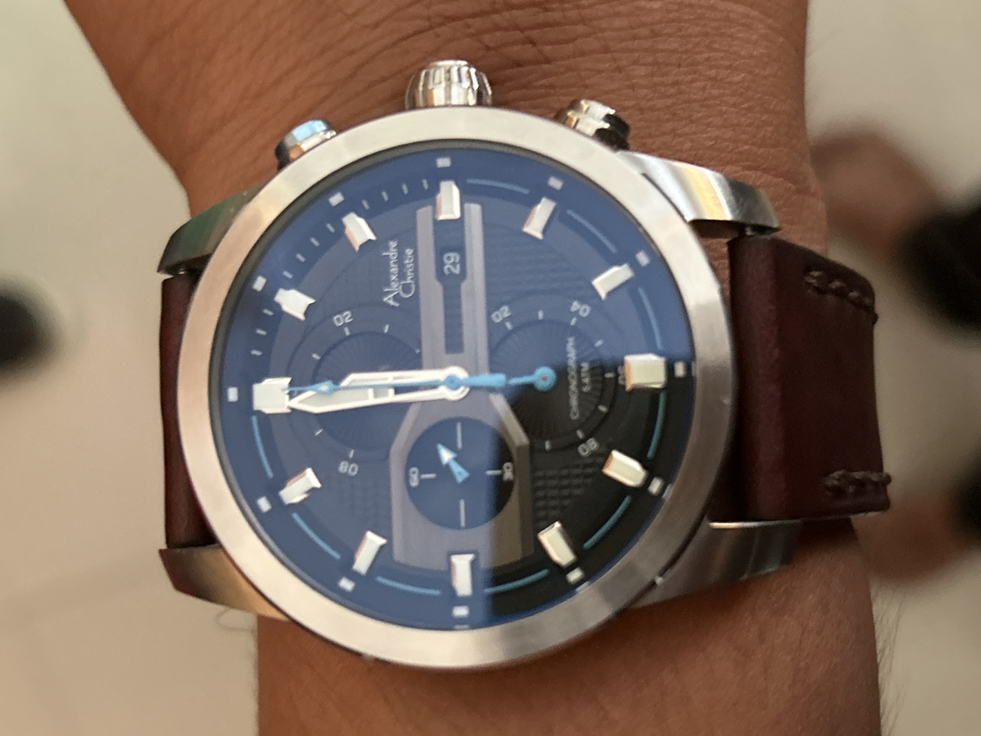 A close-up, slightly blurred photograph captures a person's wrist adorned with a metallic Alexandre Christie chronograph watch, set against a softly focused, light-colored background. The wrist displays light brown skin, suggesting an indoor setting in Lhokseumawe, Indonesia.
The watch features a round, silver-toned case and a dark brown leather strap with visible stitching. Its deep blue dial is equipped with prominent silver-colored rectangular hour markers and main hands, complemented by bright light blue secondary hands and accent lines. A date window at the 12 o'clock position clearly displays "29." Three subdials are visible: one at the top left marked "02," another at the top right also featuring "02" and "04" on its scale, and a third at the bottom with a hand pointing towards "8-."
Visible text on the watch face includes "Alexandre Christie" on the left side, and "CHRONOGRAPH 5ATM" (indicating 5 atmospheres water resistance) on the right side, just above the "08" marker. Additional numbers like "80" are also present on the dial's scales. The main hour and minute hands are positioned roughly around 10:10. The watch crown is fluted, and two pushers are visible on the right side of the case, typical for a chronograph. The image focuses sharply on the watch, rendering the surrounding details out of focus. No specific activity or emotions are discernible beyond the act of wearing the timepiece.