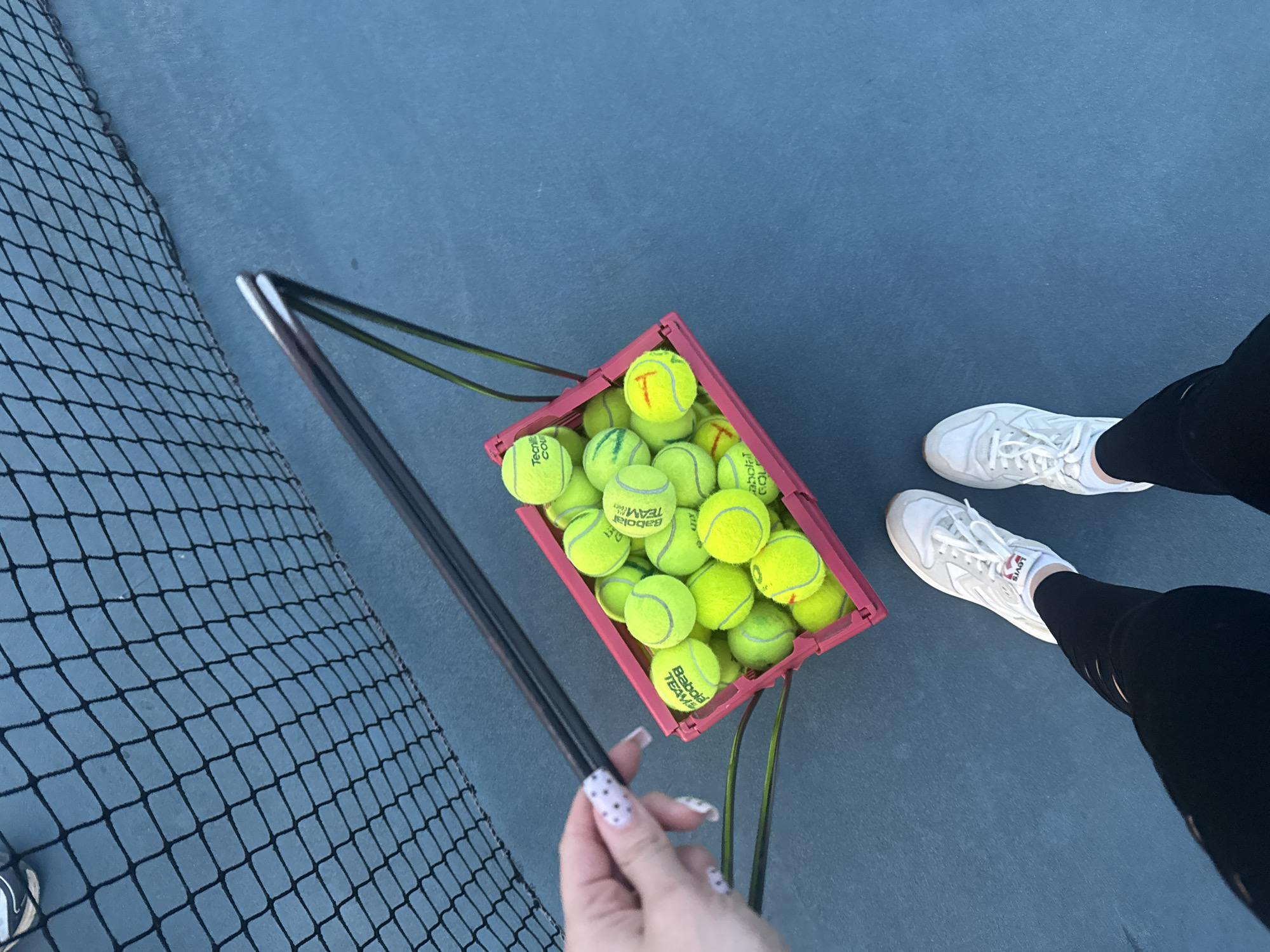 A view from above shows a section of a blue tennis court with a person standing on it. The person is wearing black leggings and white sneakers with the word "LEVIS" printed on them. The person is holding a tennis ball basket full of tennis balls with the words "BABOLAT TEAM" and "TECHNIFIBRE" on them. The handle of the basket holder is in the person's hand with polka-dot painted nails. To the left is a section of a black tennis net. Some of the tennis balls have the letter "T" on them. The location is a tennis court in Batumi, Georgia.