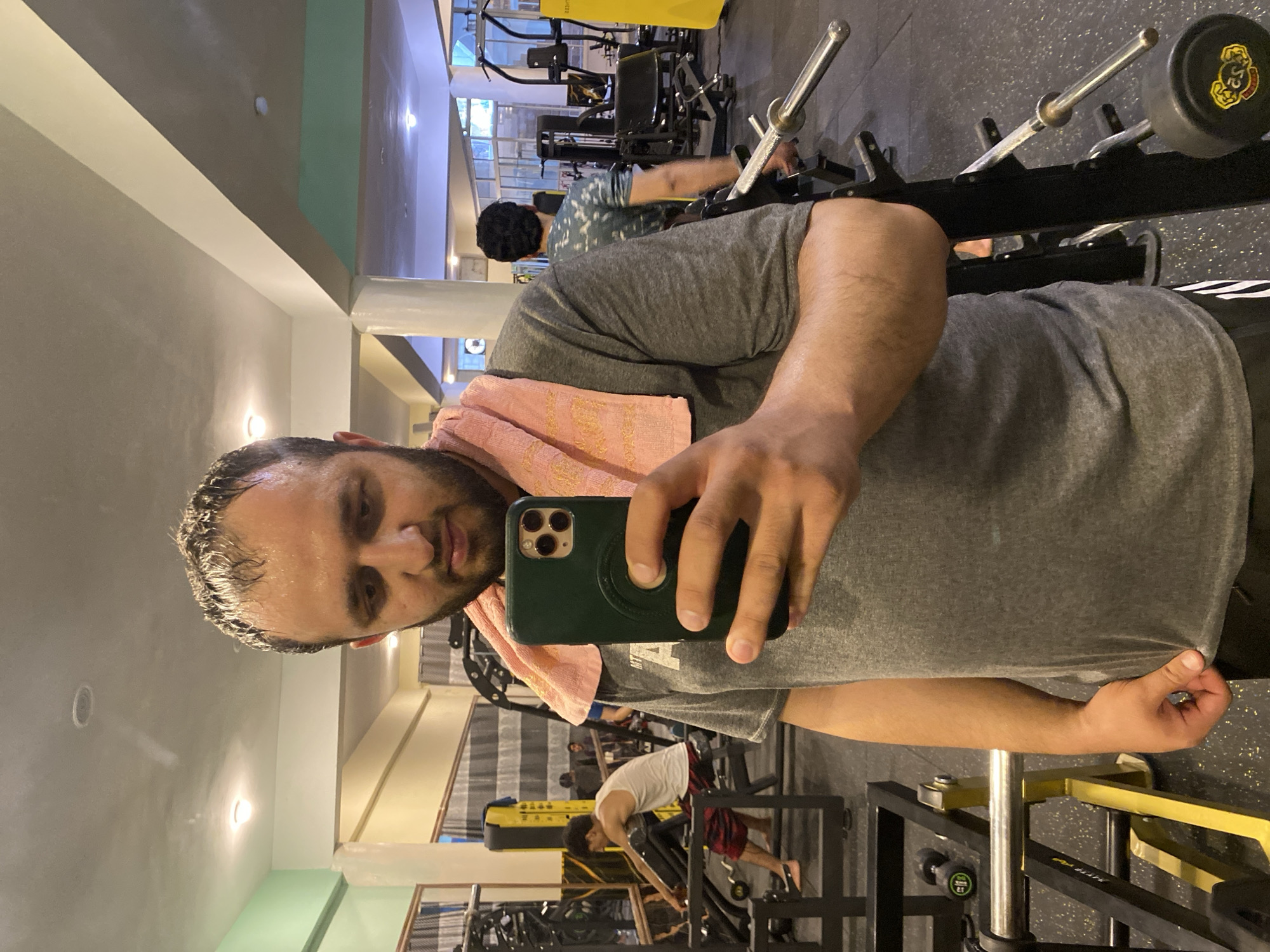 In Kandahar, Afghanistan, a man is taking a mirror selfie in a gym. He is wearing a gray t-shirt with a pink towel draped over his shoulder and is holding a green smartphone in his right hand. His hair is wet, indicating he has recently exercised. The gym is equipped with various weightlifting machines and equipment, including barbells and dumbbells, and other individuals can be seen exercising in the background. The lighting in the gym appears to be artificial, with overhead fluorescent lights.