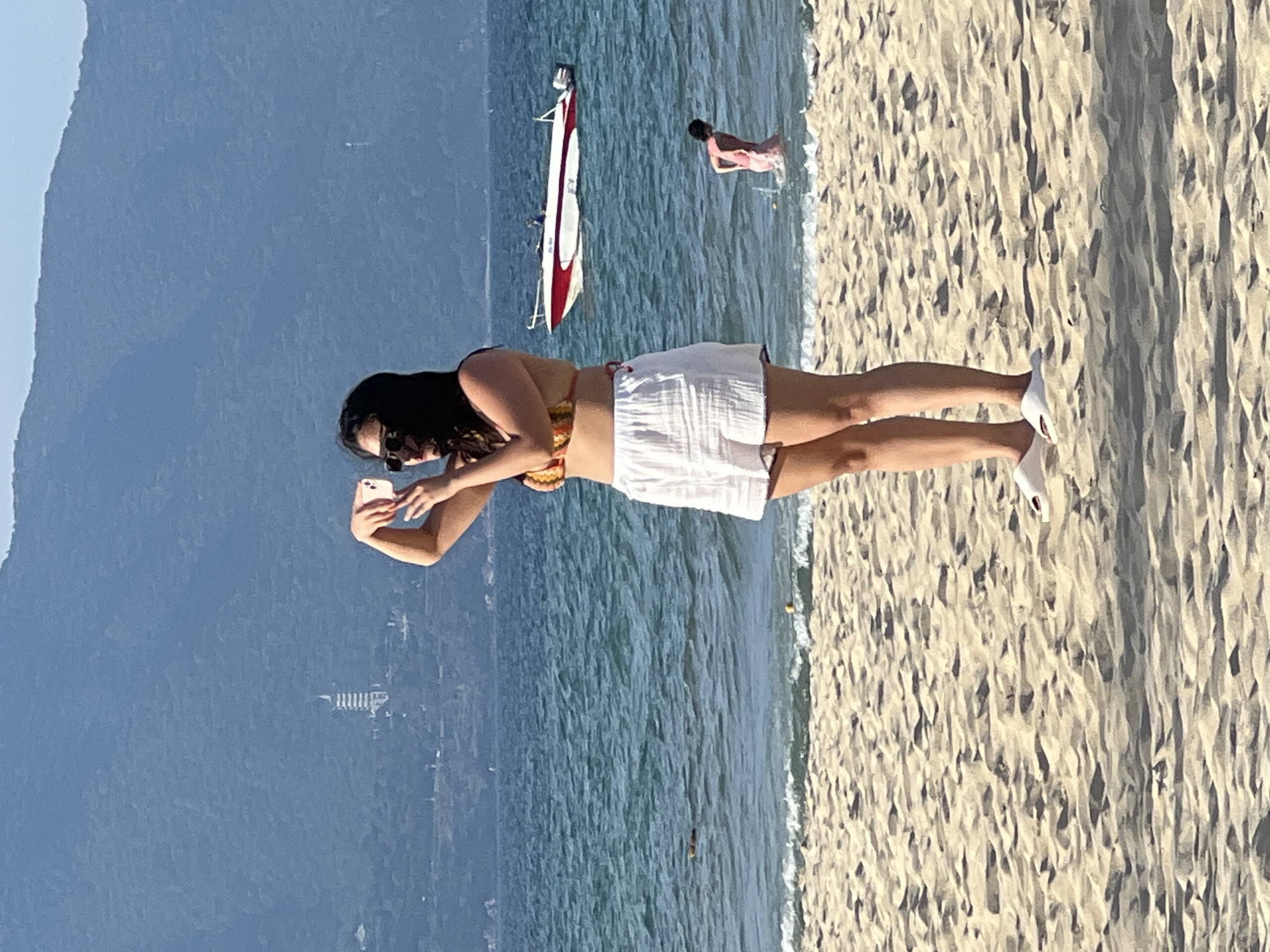 A woman stands on a sandy beach in Da Nang, Vietnam, facing the ocean and holding a smartphone to take a picture. She wears a colorful bikini top and white shorts. White slides are on her feet. The ocean is a vibrant blue with gentle waves. In the background, a red and white boat is visible on the water. Further out, a person in pink shorts wades in the ocean. Behind the water, a large, blue-green mountain range dominates the horizon. On the mountainside, a white, multi-tiered structure, possibly a pagoda, can be seen. The sky is clear and blue. The sand is light beige with visible footprints and shadows.