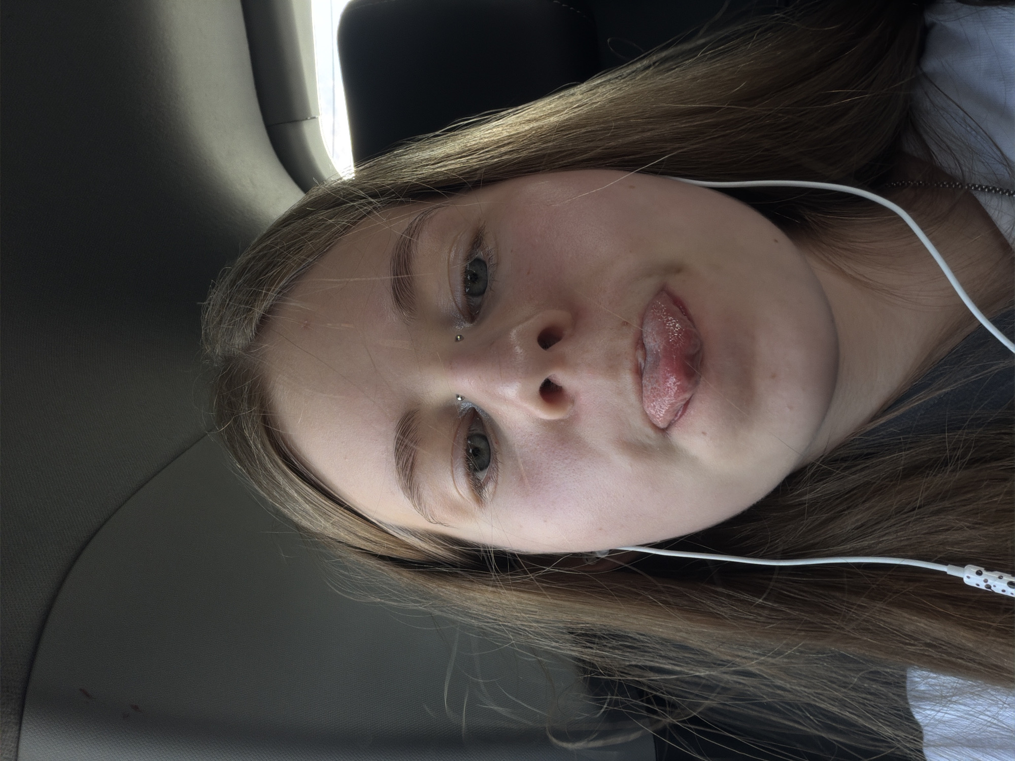 A close-up, selfie-style photograph shows a young female with long, straight, light brown hair and fair skin. She is wearing white earphones. She has a nose piercing and appears to be sticking out her tongue. Her eyes are open, and she is looking towards the camera. The background is out of focus but appears to be the interior of a vehicle, possibly a car, with a dark grey headliner visible at the top and a dark headrest in the background. There is no indication of the location being in the city of Trekhgorka, Russia.