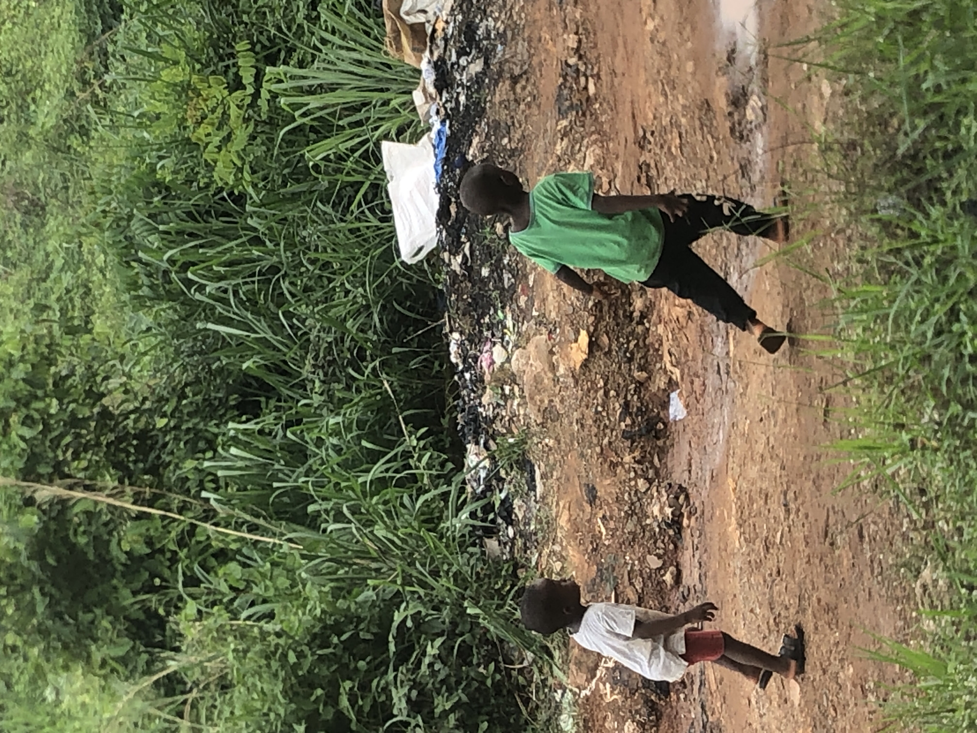 Two children are near a muddy road with debris. One child wears a green shirt and dark pants, while the other wears a white shirt and red shorts. The environment includes greenery and litter. The location is Nkwelle Ezunaka, Nigeria.