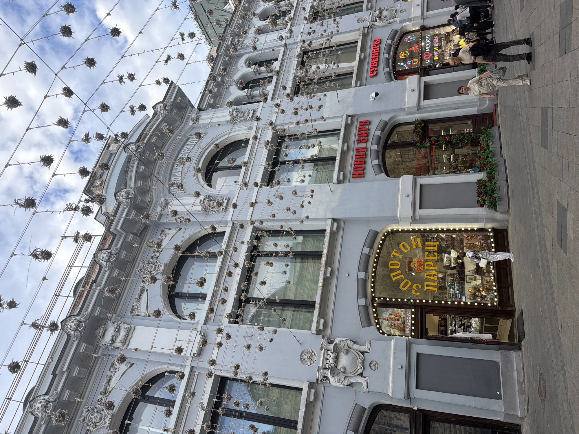 The image depicts a multi-story building exterior in Moscow, Russia. The building is adorned with architectural details, including arched windows and decorative sculptures. Strings of lights with spherical embellishments are suspended above the street. Two businesses are visible: a souvenir shop ("SOUVENIRS" in Cyrillic) and "Novaya Zara" (in Cyrillic). Several pedestrians are present, including two men walking together past a storefront. One man wearing a white outfit is standing inside the store, behind the shop window. The sky is visible in the background, with scattered clouds. The perspective of the photograph is angled upwards, accentuating the height of the building. The storefronts display a variety of items, including nesting dolls.
