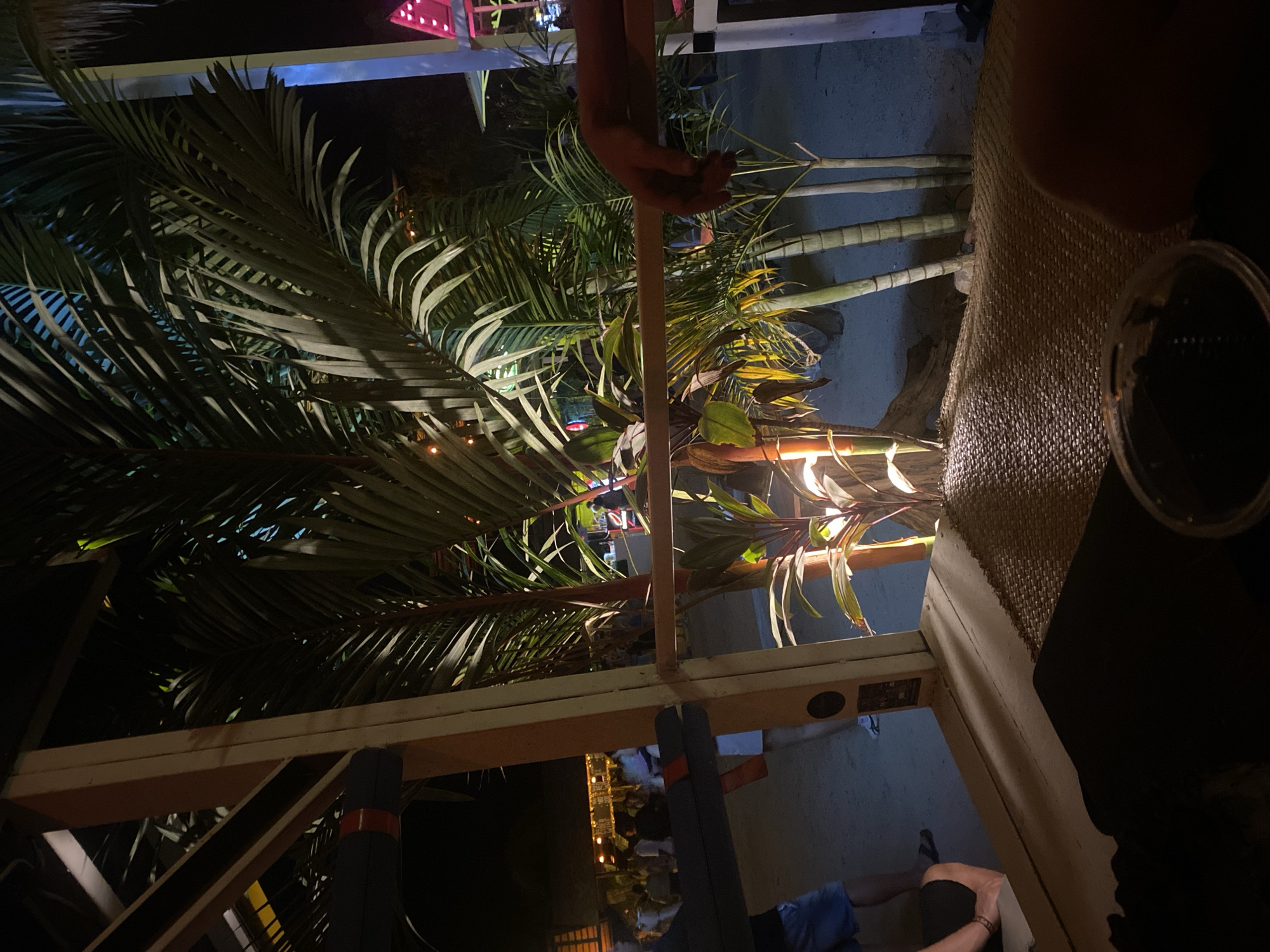 The image depicts an outdoor scene in Baan Hin Kong, Thailand, captured at night. The foreground shows part of a dining table with a woven placemat and a glass or ashtray. A person's arm and hand are partially visible above the table. In the background, there are palm trees and other tropical plants, illuminated by string lights. The scene is viewed through a partial wooden structure, possibly a pergola or building frame. The overall lighting is dim, creating shadows and highlights. The focus is primarily on the tropical vegetation and the ambient lighting.