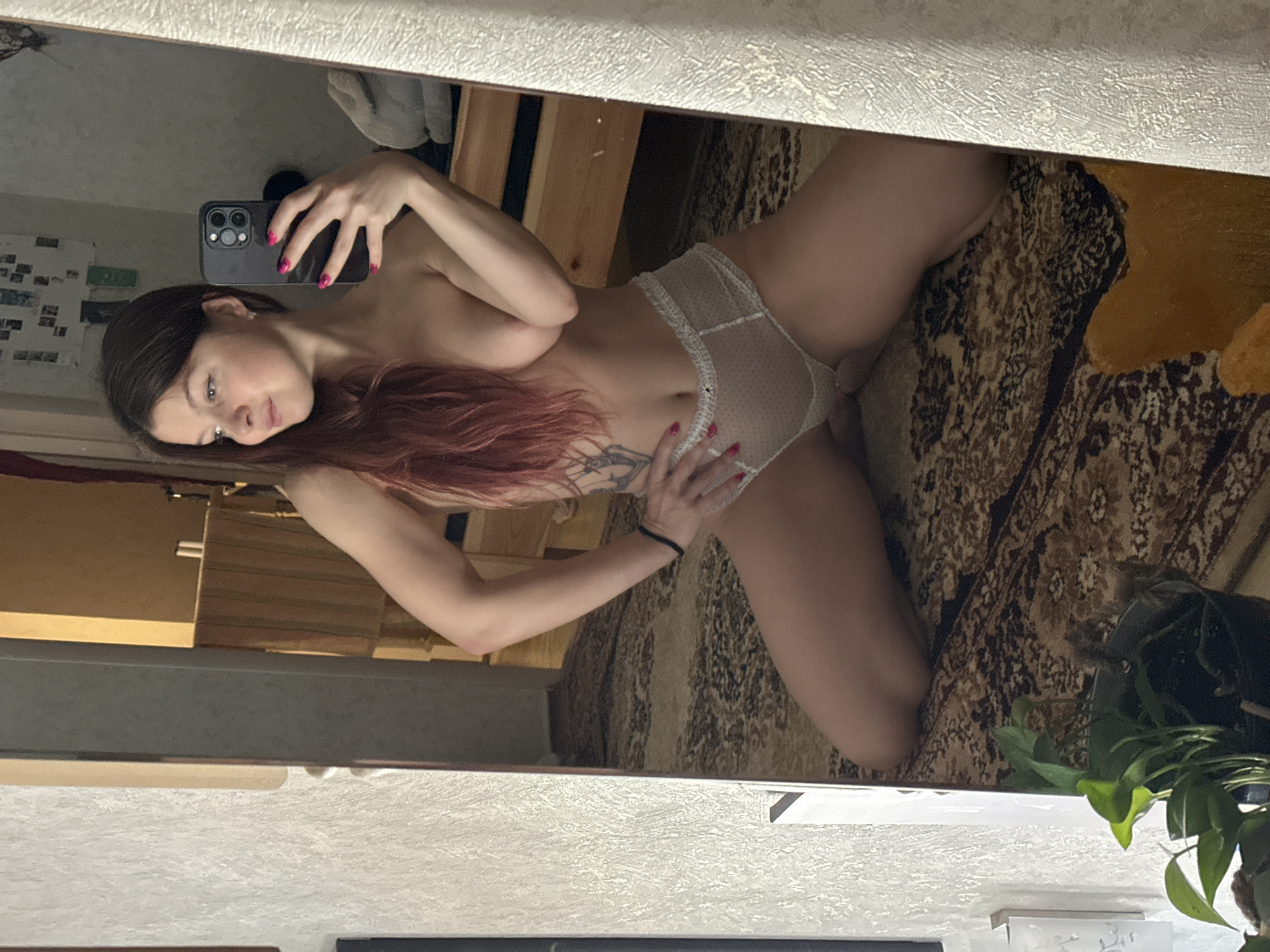 A young woman is captured in a mirror selfie, appearing to be kneeling or posing on a patterned rug. She is wearing white, sheer, polka-dotted underwear. Her body is angled, with one hand resting on her hip and the other holding a dark-colored smartphone, used to take the picture in the mirror. Her fingernails are painted bright pink. She has long, dark hair with visible reddish highlights, and a tattoo is visible on her left torso. Her gaze is directed towards the phone, with a neutral expression.
The setting is indoors, reflected in the mirror. Behind her, a wall with a light, textured surface is visible. Parts of wooden furniture, possibly a bed frame or shelving, are seen to the left in the reflection. A patterned rug in shades of brown, beige, and black covers the floor. In the bottom right of the reflection, a potted plant with green leaves is partially visible. The lighting is diffused and indoor. The image is rotated 90 degrees clockwise from its natural orientation. No visible text is present in the image.