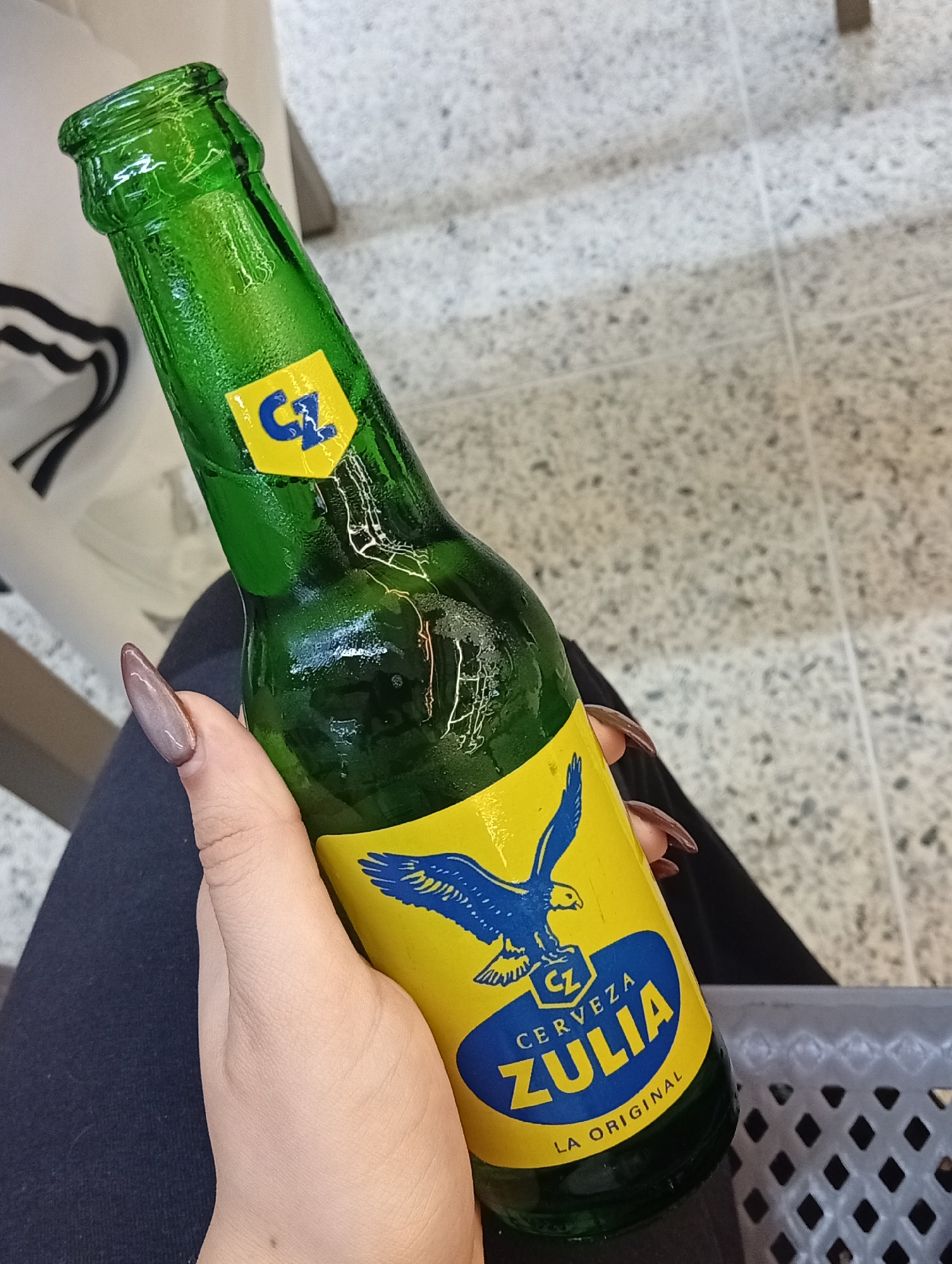 A person is holding a green glass bottle of Zulia beer, a Venezuelan brand. The bottle has a yellow label with a blue eagle and the words "Cerveza Zulia" in blue lettering. The bottle is slightly wet, indicating it might be cold. The person is wearing dark blue pants. The fingernails on the hand holding the bottle are long and almond-shaped, with a brown, cat-eye polish. A grey plastic lattice basket is partially visible in the background to the right. The setting appears to be indoors, possibly a store or a home. There is no evidence to suggest a specific location in Porlamar, Venezuela.