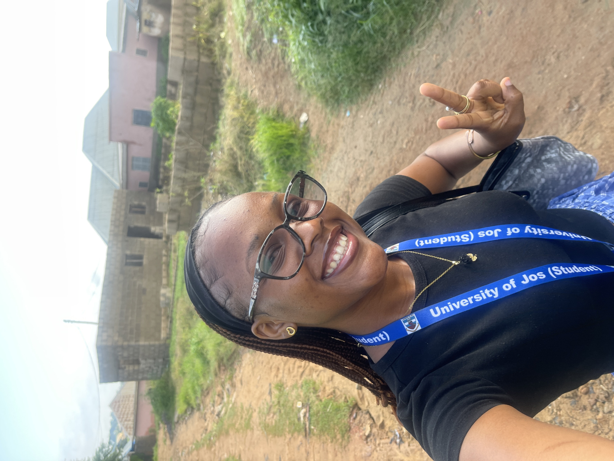 A young woman with braided hair and glasses smiles at the camera, holding up two fingers in a peace sign. She is wearing a black t-shirt and a blue lanyard around her neck that reads "University of Jos (Student)". She is standing in a dusty outdoor environment with buildings and vegetation in the background. The location is identified as Rafin Par, Nigeria.