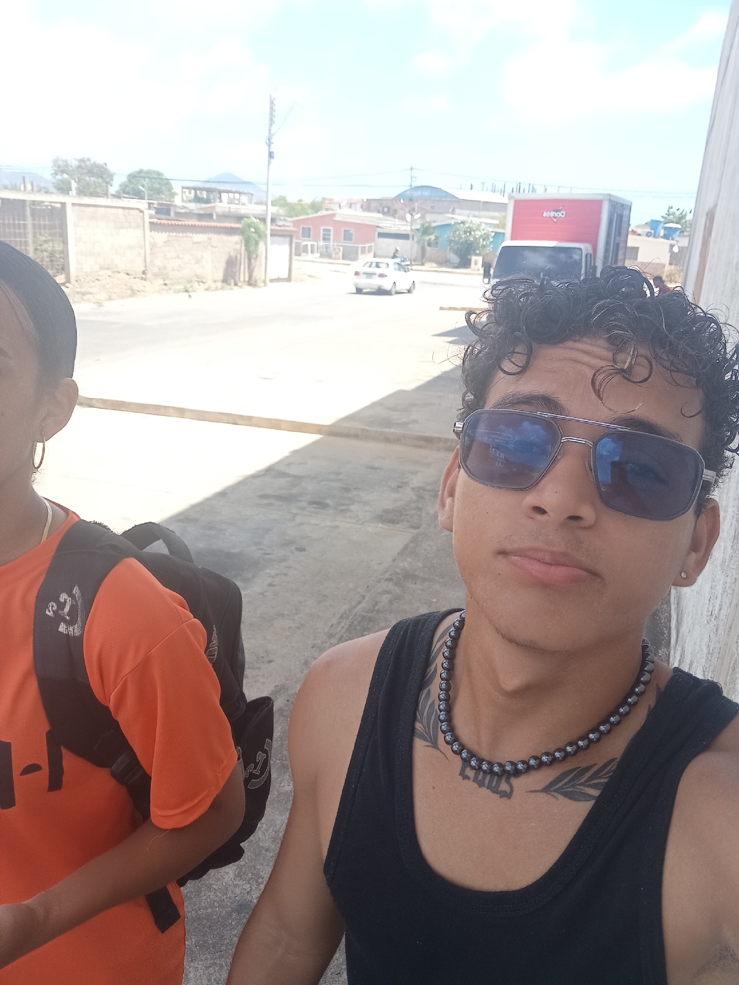 In the city of Juan Griego, Venezuela, two young individuals, a male and a female, are captured in a selfie outdoors. The male, positioned in the foreground and facing the camera, wears a black tank top and a beaded necklace. He sports sunglasses and has curly dark hair. A tattoo is visible on his chest. The female, partially visible to the left, wears an orange t-shirt and has a backpack on. The background reveals a street scene with buildings, a red truck, and a white car. The overall setting appears to be a sunny day in an urban environment.