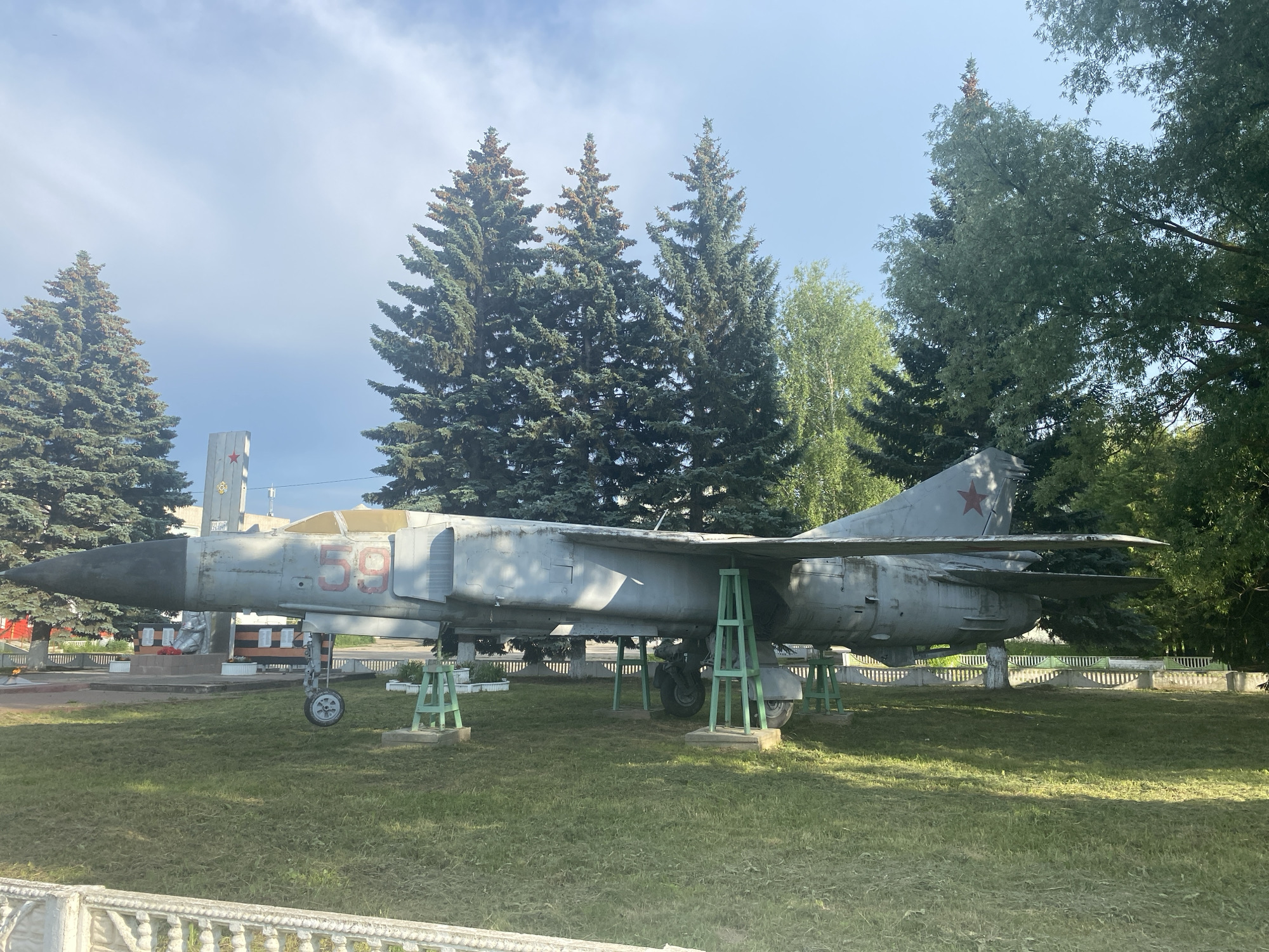 An old, grey Soviet-era fighter jet, marked with the number "59" in red on its fuselage, is displayed outdoors on a grassy field in the city of Aisha, Russia. The aircraft is supported by green metal stands. The jet has a pointed nose, swept wings, and a red star insignia on its tail fin. In the background, tall evergreen trees and other deciduous trees form a backdrop under a partly cloudy blue sky. A white decorative fence runs along the front of the grassy area. To the left of the jet, a monument with a statue and war memorial elements is visible, including a flame and several display boards.