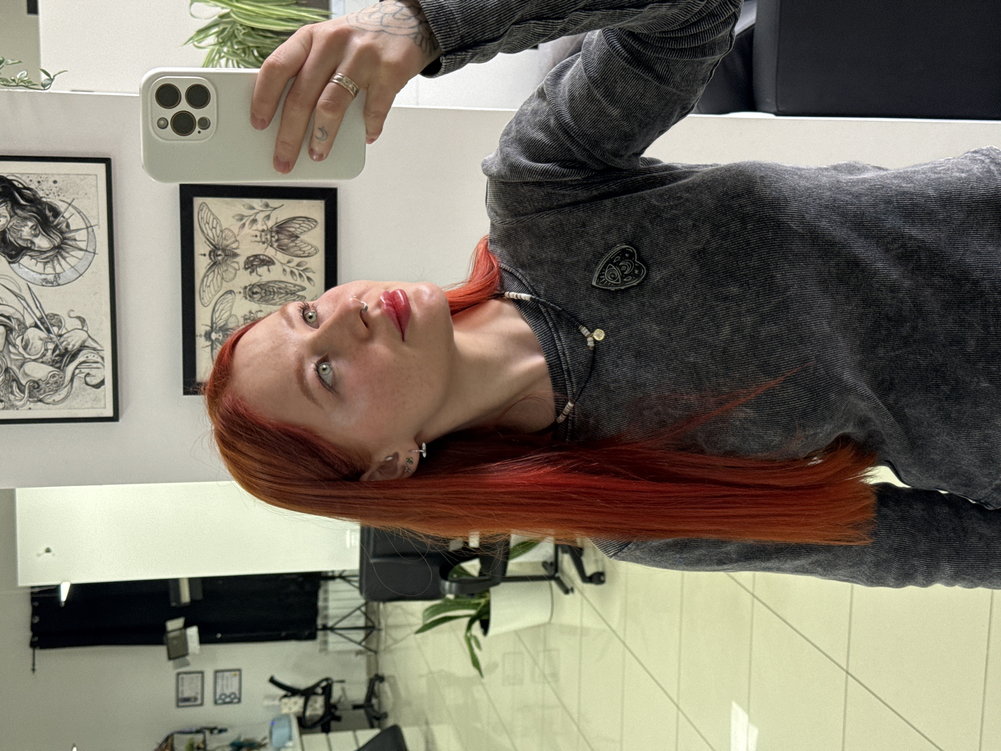 A young, fair-skinned woman with red hair is taking a selfie with a white smartphone. She has multiple piercings in her ears and nose. She is wearing a grey corduroy top with a black and white eye-shaped pin. Her right hand, holding the phone, has tattoos on the wrist and fingers. The background includes two framed artworks on a white wall, one depicting a woman and a moon, and the other displaying illustrations of insects. There is also a potted plant on a shelf. The overall environment appears to be a well-lit interior space. The presence of a tattoo chair, lighting equipment, and additional framed artwork suggests the location may be a tattoo studio or art gallery. There is no indication of the city of Rostov-on-Don, Russia.