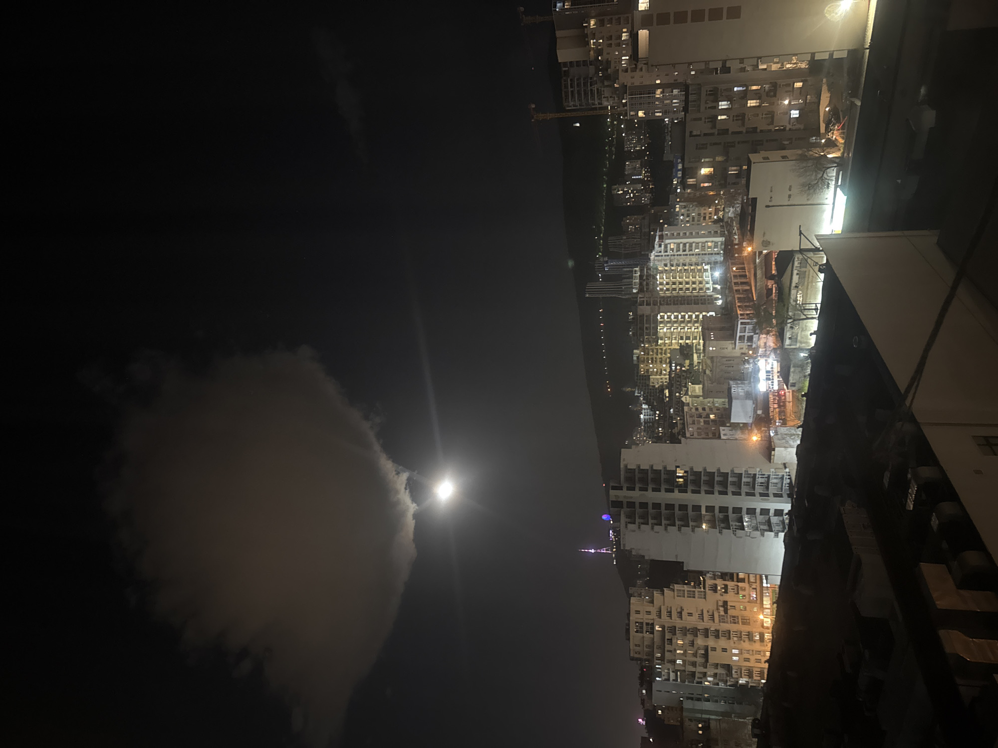 The image presents a night view of Tbilisi, Georgia. The lower-left quadrant of the image is dominated by a large, bright cloud partially obscuring a full moon. To the right of the cloud, the city's skyline is visible, illuminated by artificial lights from buildings and street lamps. The silhouette of a high-rise building or structure stands out against the sky. The image is taken from an elevated position, providing a wide view of the city. The urban environment is characterized by numerous multi-story buildings, indicating a densely populated area. The overall atmosphere is dark, with light sources concentrated in the city's built environment.