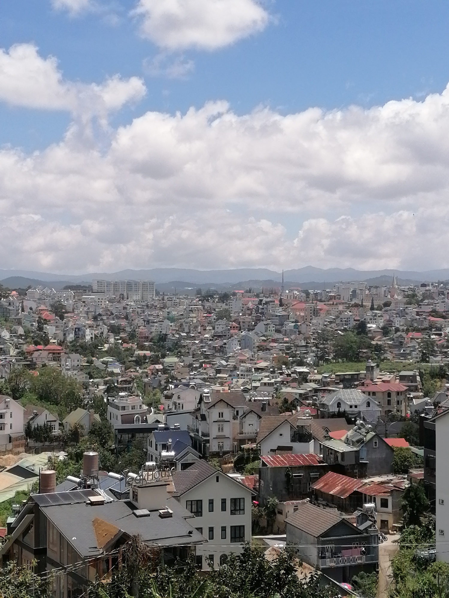 A high-angle, full shot captures an expansive cityscape of Da Lat, Vietnam, under a bright, partially cloudy sky. The urban sprawl extends into the distance, characterized by a dense collection of houses and buildings with varied architectural styles and roof colors, including red, gray, and white. The terrain appears hilly, with structures built on slopes and a significant amount of greenery interspersed throughout the residential areas. In the foreground, a few houses are more distinctly visible, one with a dark roof featuring solar panels and two large water tanks. The distant horizon is marked by a range of mountains. The overall impression is that of a densely populated city nestled within a natural, hilly landscape.
