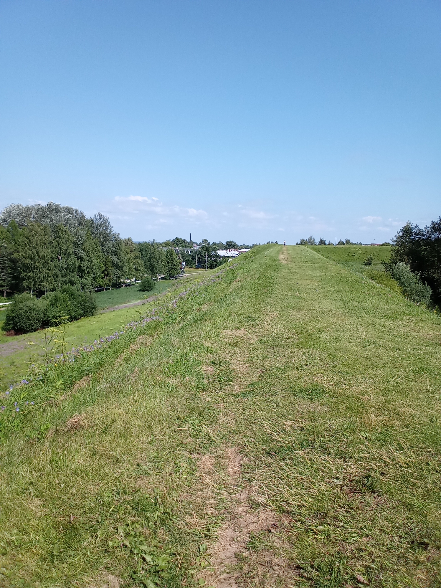 The image depicts a sun-drenched day with a clear blue sky. A grassy embankment, or dam, slopes upwards towards the horizon. A dirt path winds its way along the crest of this embankment. To the left of the embankment, a line of lush green trees forms a backdrop, with scattered buildings visible beyond the trees. In the distance, a lone figure walks along the path at the very top of the embankment. The overall scene suggests a park or natural area within or adjacent to the city of Belozersk, Russia, likely part of its historical fortifications or landscape.