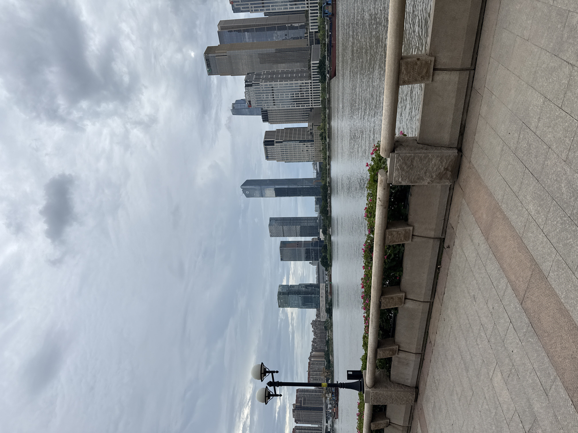 A wide, tranquil river flows through a city on a cloudy day. On the far bank, a dense collection of modern skyscrapers with varied architectural styles dominates the skyline, reflecting the overcast sky. Several buildings are tall and slender, while others are broader and more angular. A large boat, appearing to be a cargo vessel, is visible in the middle of the river, heading towards the right side of the frame. The foreground features a paved walkway lined with a stone wall and decorative planters containing green foliage and small pink flowers. A single lamppost with two spherical lights stands near the left edge of the walkway. The scene appears to be a public promenade or riverfront area in Haizhu District, China, offering a view of the city's contemporary urban landscape against a dramatic, cloudy sky.