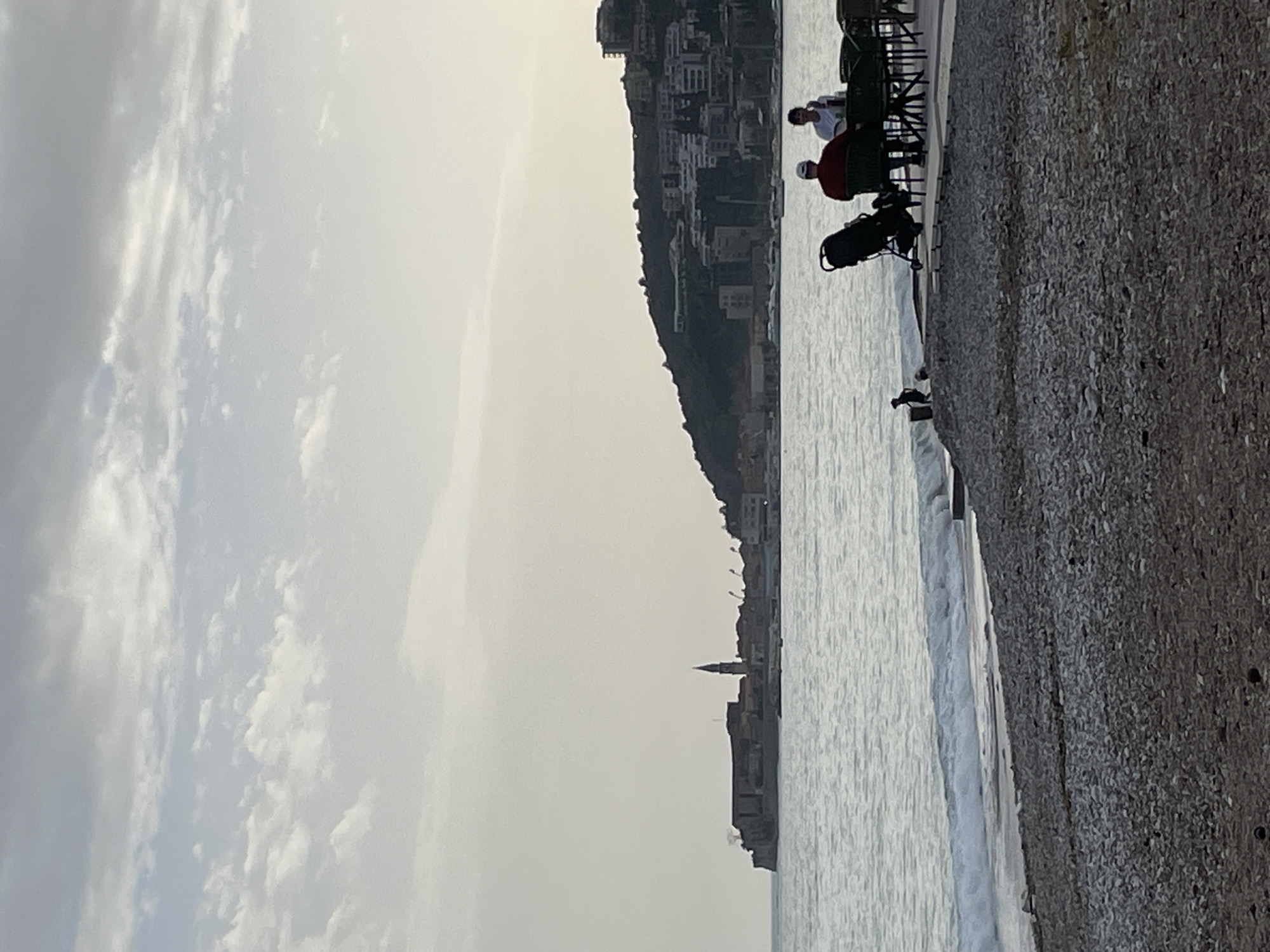 The image shows a scene in Budva, Montenegro. A stone embankment lines the right edge of the frame, while the sea stretches to the left. In the distance, a settlement with buildings and a church spire is visible. Two individuals are seated at an outdoor dining area with green chairs, possibly a restaurant or cafe. A black stroller sits nearby. The sky is overcast with clouds. The water appears calm with small waves.