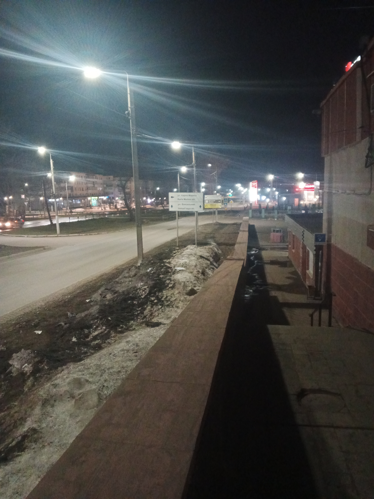The image depicts a nighttime scene in Chaikovsky, Russia. In the foreground is a wall bordering a raised sidewalk area leading to a building on the right. The building appears to be two-toned, with a lighter colored base and a red brick upper section. The raised sidewalk area is dark and shadowed. Adjacent to the sidewalk wall is a strip of land with melting snow and debris. A road runs parallel to this strip of land. Streetlights illuminate the road and surrounding area. In the distance, buildings and signs, some brightly lit, are visible along the street. A sign with direction information is partially visible; the sign indicates that the street to the left is Karla Marksa st. and the street forward is Vokzal naya st. Several lights appear to be near an eatery in the background. A vehicle is visible traveling along the road.