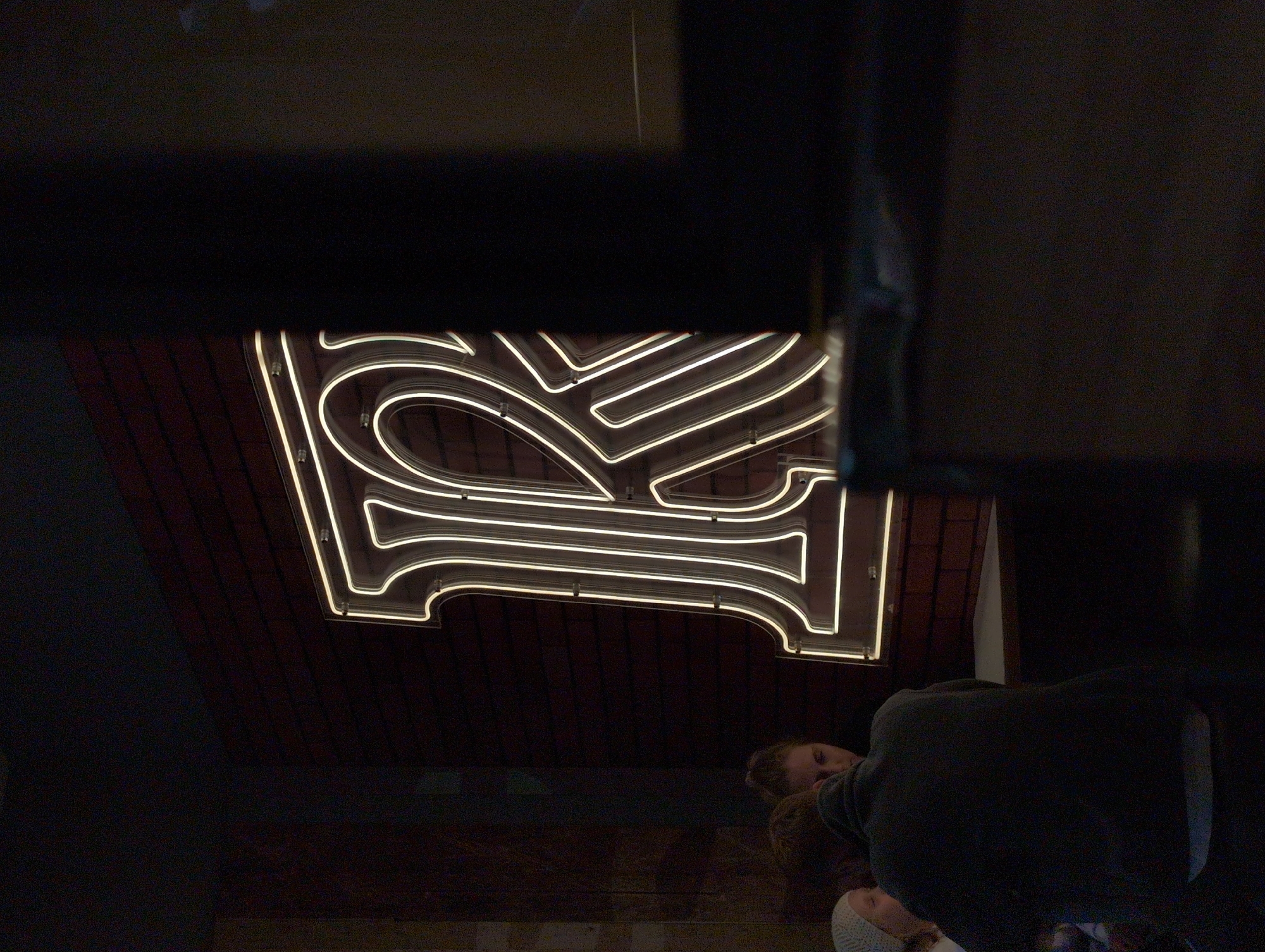 The image shows a low-angle view of a neon sign with a stylized letter "I" against a dark brick background. The sign is brightly illuminated with a white glow. At the bottom of the frame, parts of people are visible; possibly two individuals are near the sign. A person wearing a dark gray top is present, and another person with fair skin and light hair is visible behind. Part of the image is obscured by the frame in the foreground.
The location is the city of Izhevsk, Russia.