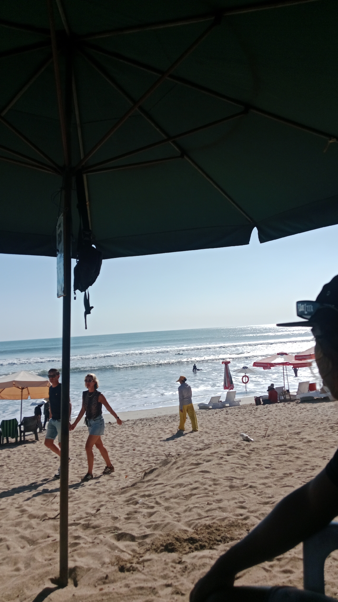 This is a medium-angle shot of a beach in Kuta, Indonesia, captured from under a large, dark green beach umbrella. The sand is light brown and textured, with some small dunes in the foreground. In the mid-ground, a man and a woman are walking on the beach. The man is wearing a dark t-shirt and light-colored shorts, and sunglasses. The woman is wearing a patterned top and denim shorts, and sunglasses. They are walking towards the viewer, slightly to the left.
Further back, near the water's edge, a person in a yellow shirt and yellow pants stands facing away from the viewer. Several beach chairs and umbrellas are visible, some red and white. The ocean is calm with gentle waves breaking on the shore. There are a few people in the water, and a surfer is visible in the distance. The sky is clear and blue. A black bag hangs from the pole of the umbrella in the foreground. The perspective is from someone seated or standing beneath the umbrella, looking out towards the beach and ocean.