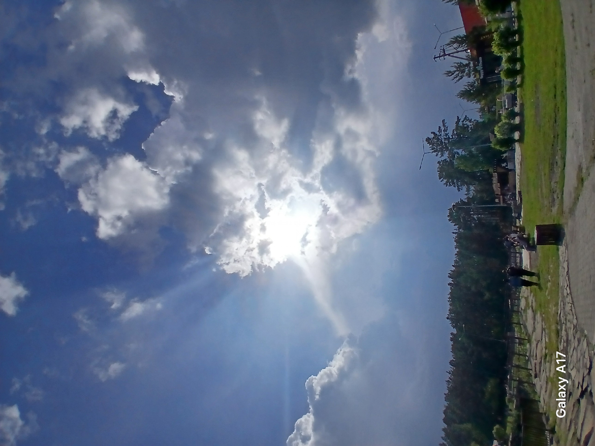The image, captured in Yekaterinburg, Russia, depicts an outdoor scene under a sky with significant cloud cover and prominent crepuscular rays. Bright sunlight penetrates through a central cloud formation, casting distinct beams downwards. The upper left portion of the sky is clearer blue, while the right and lower sky exhibit darker, denser cloud masses.
In the foreground, a paved path with irregular stone shapes extends from the lower left, curving towards the right. To the right of the path is a grassy area. A brown cylindrical object, possibly a waste bin, is positioned near the path on the right side of the lower frame. Two individuals are walking on the path in the mid-left portion of the lower half, moving away from the viewer. One individual wears a blue top, and the other a darker garment.
Beyond the path and grass, a fence or railing structure is partially visible. A dense line of dark green trees, primarily conifers, forms a backdrop across the image's width. To the right of the walking individuals, on a slightly elevated grassy area, multiple people are observable, with some seated on benches. A row of small, manicured green bushes runs along this area.
In the background, behind the trees, multiple tall streetlights or utility poles are distributed. Structures are visible, including one on the far right with a red facade and another appearing to be a commercial establishment with visible text, "Happy Place," on its signage. The image bears a "Galaxy A17" watermark in the bottom left corner.