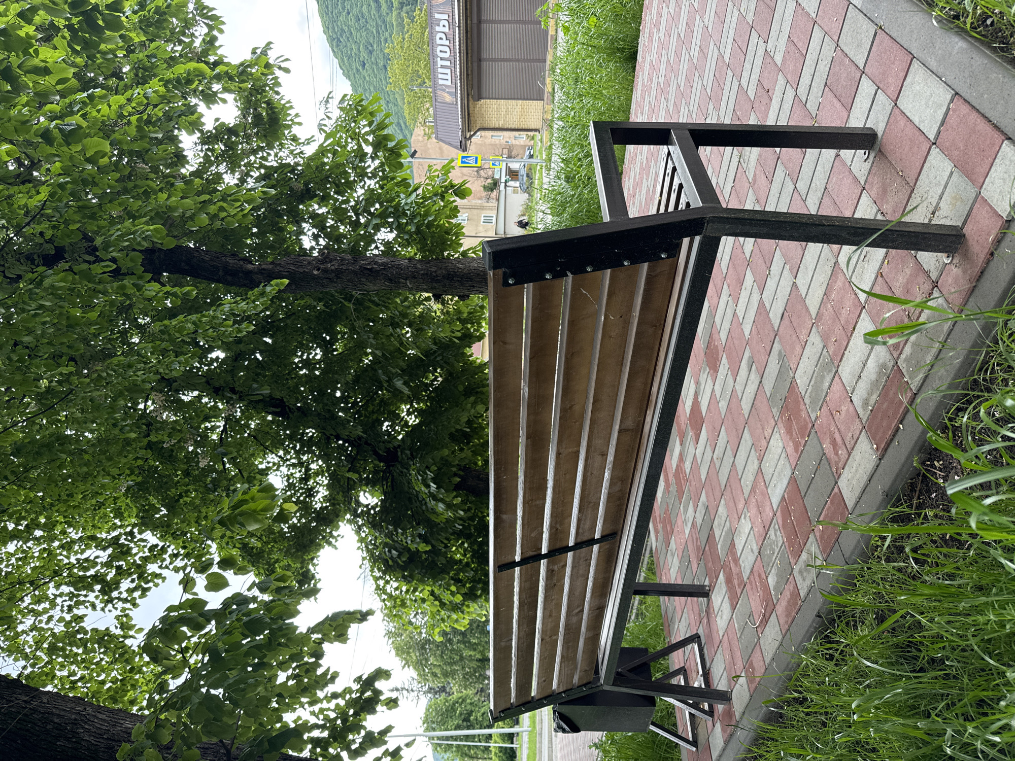 A park bench constructed with a metal frame and wooden slats sits adjacent to a tree and a paved walkway. The walkway is composed of red and gray rectangular pavers arranged in a patterned design. Green grass grows on the outer edge of the walkway. In the background, buildings are visible, including one with a sign in Russian. The overall scene suggests a public park or rest area in Karachayevsk, Russia.