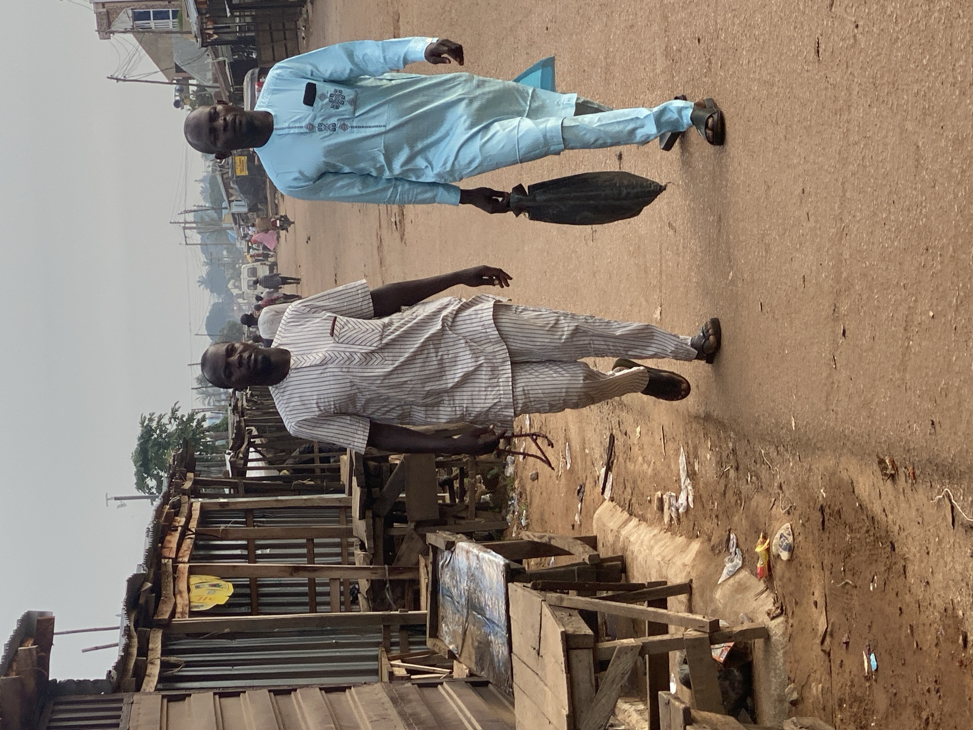 Two men walk on a dusty, reddish-brown road in Sabon Tasha, Nigeria. The man on the left wears a white and grey striped short-sleeved tunic and matching trousers. He carries a pair of eyeglasses in his right hand. The man on the right wears a light blue long-sleeved tunic and matching trousers. He carries a dark, elongated bag in his right hand. Both men are wearing sandals. They are walking away from the camera, towards the center of the frame.
To the left of the road are several makeshift wooden stalls with corrugated metal roofs, some of which appear to be empty or under construction. Tables and wooden structures are visible in front of the stalls. There is debris and litter on the ground, including plastic bags and paper.
In the background, the road extends into the distance, with more buildings and structures visible on either side. A few other people are walking in the distance, and a motorcycle is also visible. The sky is overcast. The overall scene depicts a street in a developing urban area.
