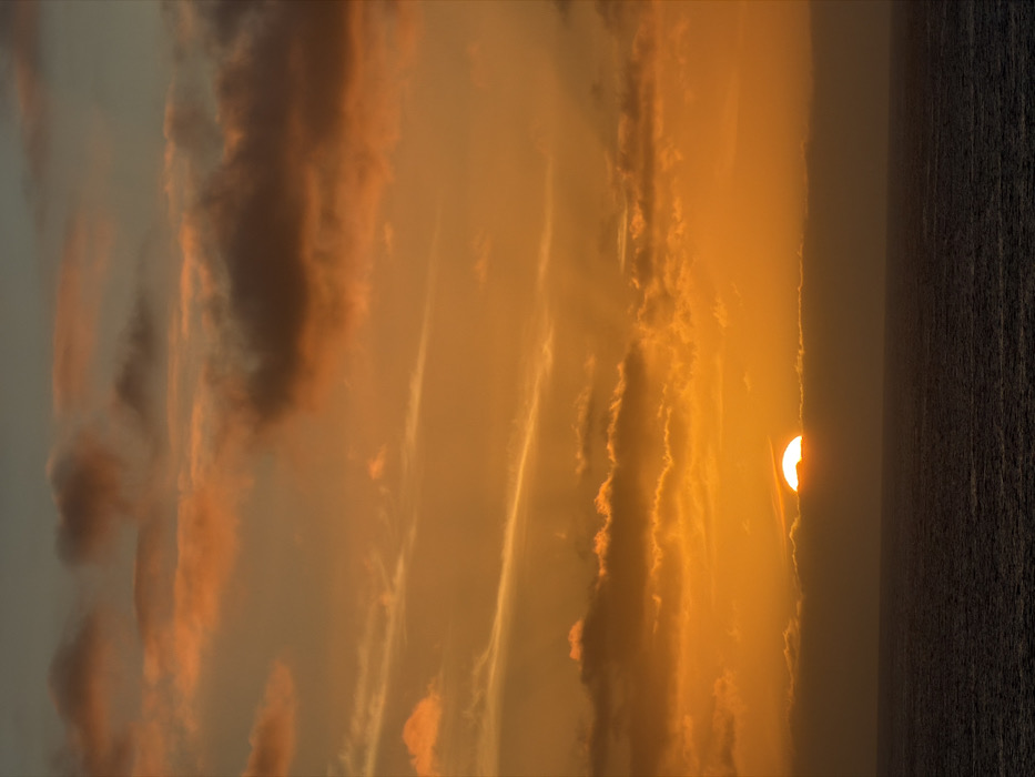 An upside-down vertical image shows a sunset over the ocean. The sun is visible on the right, partially obscured by thin clouds. The sky is filled with orange and yellow hues, transitioning to grey on the left. Dark, thin clouds stretch vertically throughout the frame, adding texture to the sky. The sea on the right is dark and appears calm.