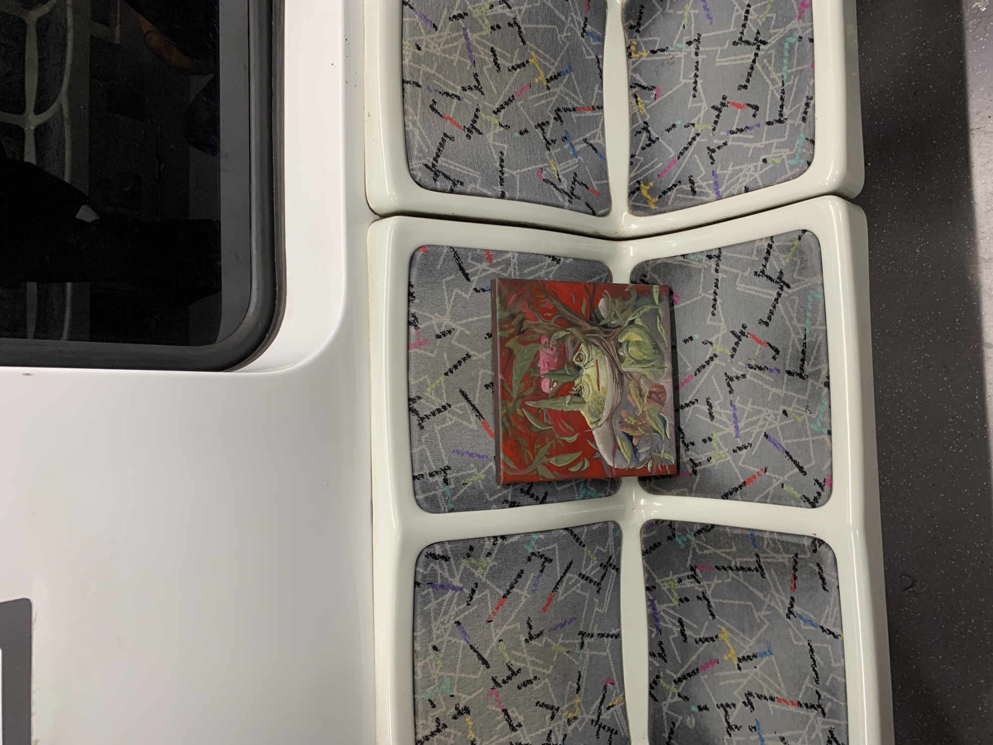 A small painting has been placed on the back of a public transit seat. The seat has a patterned grey fabric. The background is red, and it has a partial floral motif. The public transit vehicle is near Buenos Aires, Argentina.