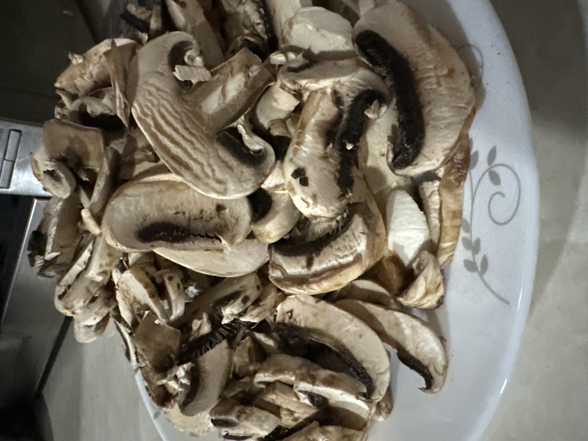 A plate of sliced mushrooms is presented in a close-up shot. The mushrooms are predominantly brown and white, with visible gills on the underside of some slices. They are piled on a white plate that features a subtle decorative pattern of swirling lines and small leaf shapes. The background is blurred, but appears to be an indoor setting with hints of metallic surfaces and a countertop. There is no visible indication of the city of Sasbaga, Uzbekistan.