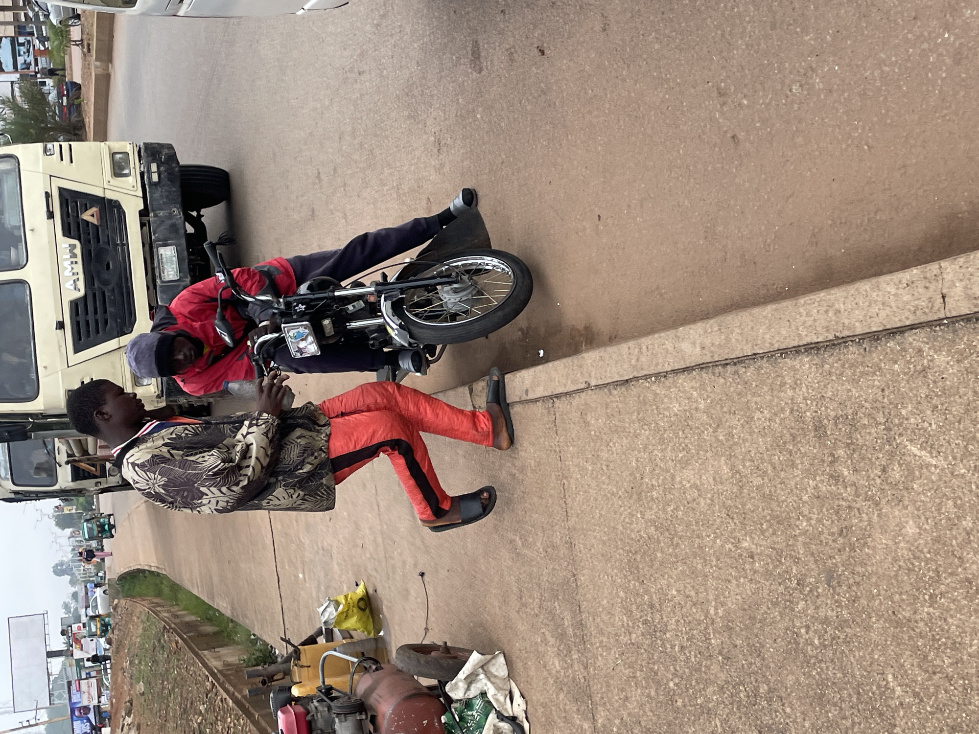 Two young men are interacting with a motorcycle on a paved road, likely in Jos, Nigeria, based on context from related images. The man on the left is wearing a patterned jacket and bright red trousers with black stripes. He is holding a plastic bottle and appears to be leaning over the motorcycle, possibly explaining something or handing an item to the other man. The man on the right is seated on the motorcycle, wearing a red jacket and dark trousers. He is looking down at the motorcycle's handlebars and has a dark cap on his head. The motorcycle is black and silver with a visible front wheel and license plate. In the background, a large white truck with "AMW" written on the front is parked on the right side of the road. The environment appears to be an urban setting with a sidewalk on the left and other vehicles and structures visible in the distance, including a billboard. There are also some items on the ground near the man on the left, including a red air compressor and some yellow and white bags. The road surface is paved with concrete.
