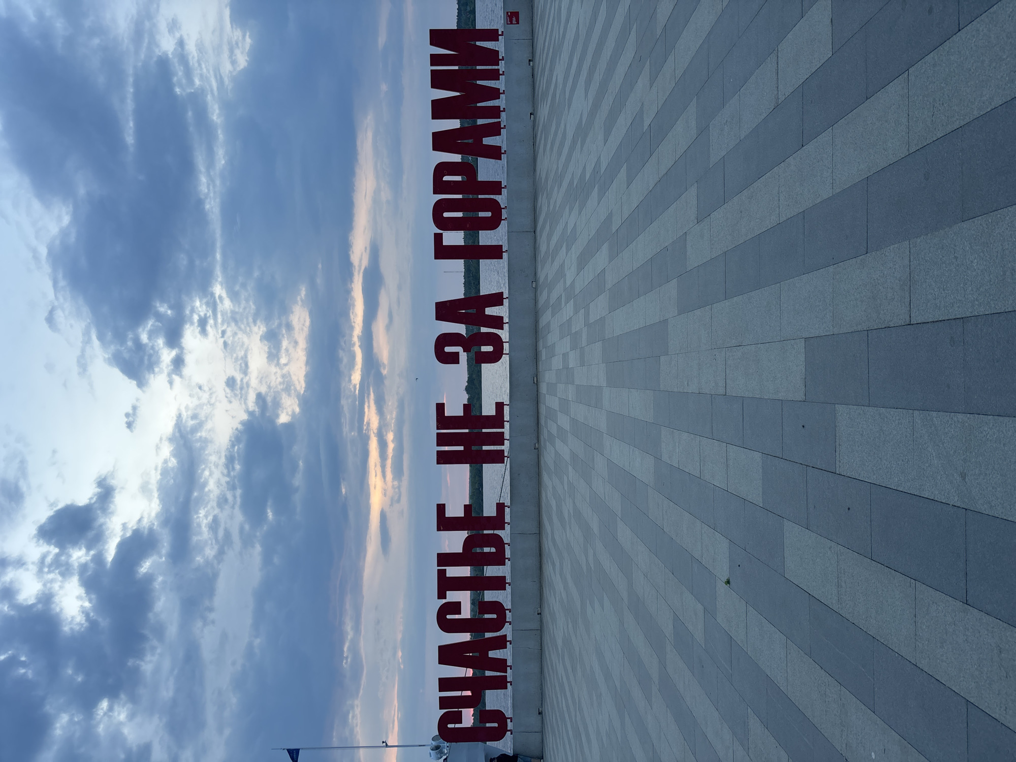 The image shows a horizontal public art installation in Perm, Russia. The red Cyrillic letters, "Счастье не за горами" (Happiness is not far off), are located on a grey-paved walkway along the Kama River embankment. The sky above is cloudy, with a hint of sunset colors. The water of the river is visible in the background between the letters.