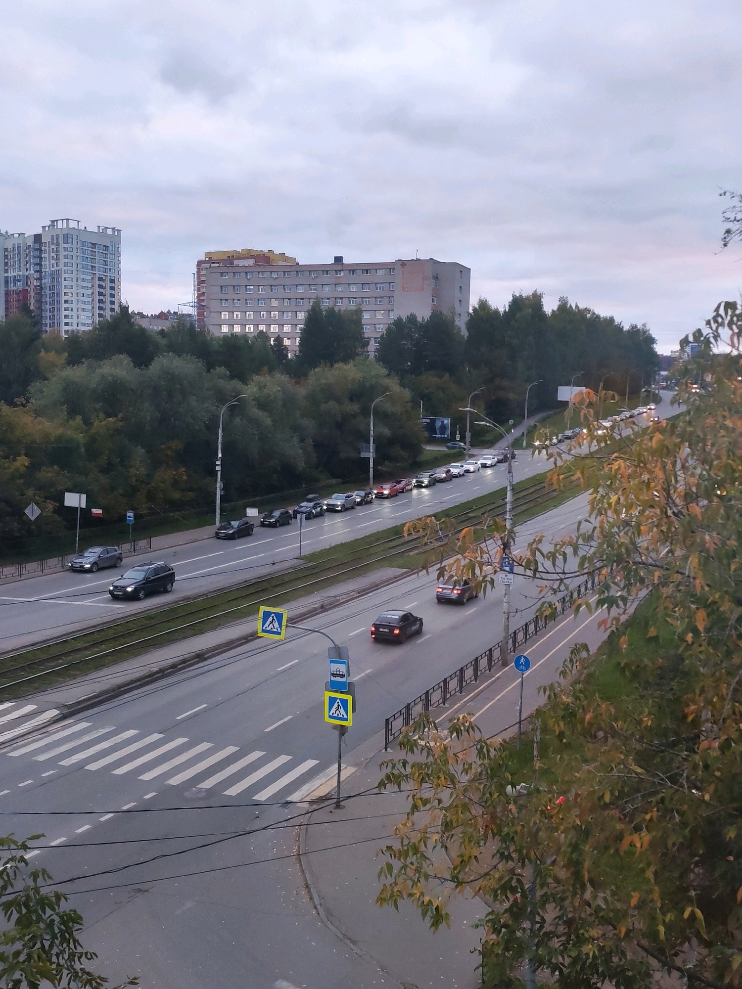 The image captures an overcast day in Izhevsk, Russia, on Kommunarov Street. In the foreground, a multi-lane road is visible with cars driving and parked. A pedestrian crossing with white stripes is marked on the road. Traffic lights and pedestrian crossing signs are present on the roadside. On the left side of the road, tram tracks are visible. In the background, several multi-story buildings and a dense line of trees can be seen. The sky is cloudy, and the overall lighting suggests it is either early morning or late afternoon.