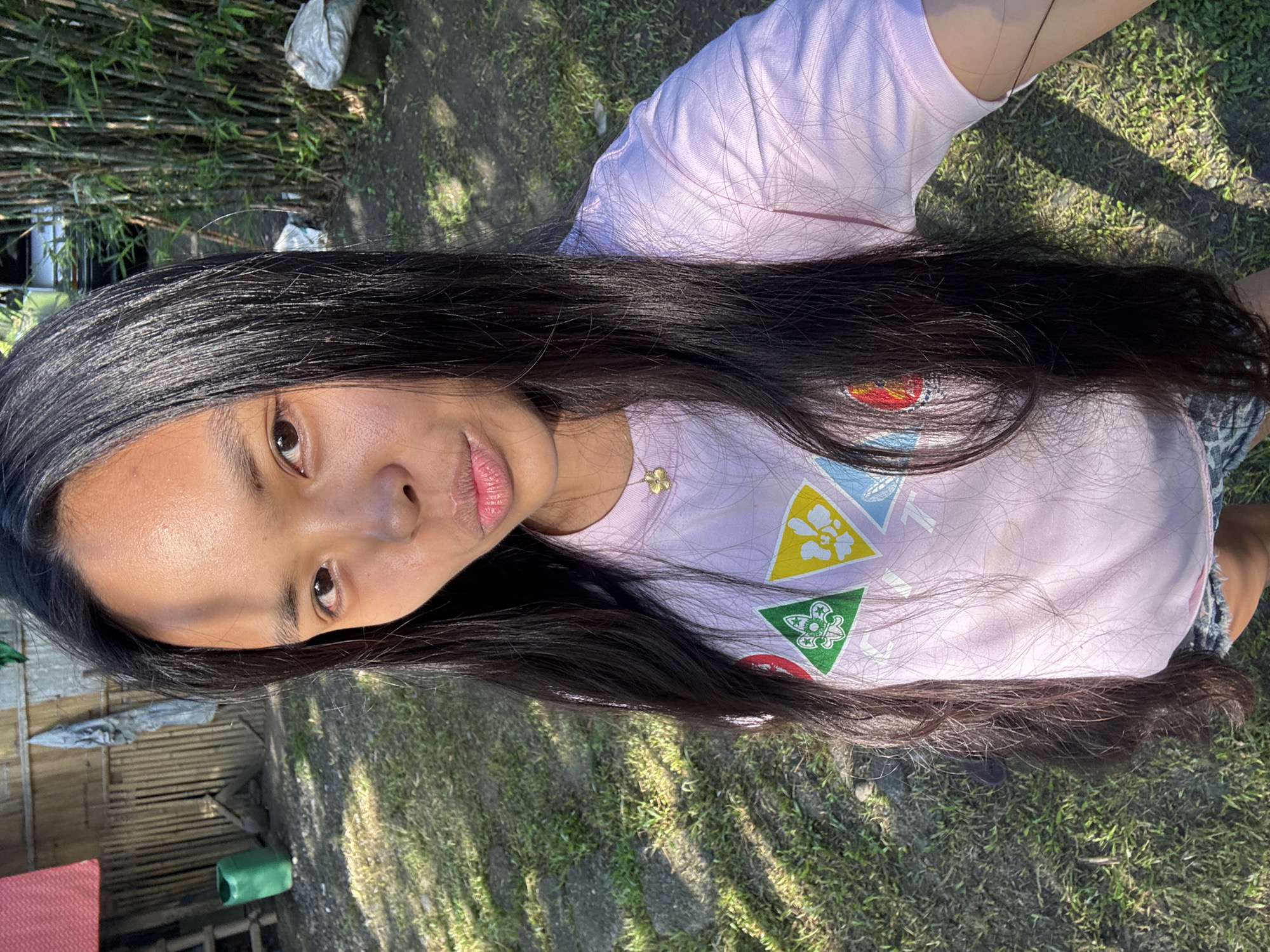 An East Asian female subject with long, straight, dark hair and dark eyes is depicted in a close-up outdoor shot, presumably in Carmen, Philippines. She is looking directly at the camera with a slight pout. Her skin shows a radiant sheen, with sun glare visible on her forehead. She is wearing a light pink short-sleeved t-shirt adorned with multiple triangular emblems; one is green with a white trefoil symbol, and another is yellow with a white floral design. A gold-colored necklace with a small flower pendant is visible around her neck. Her left arm is slightly raised, suggesting she is holding the camera. The background to her left features a rustic bamboo wall, a red fabric item, and a green plastic jug on the ground. To her right, a dense stand of bamboo stalks is visible, with hints of a dark-colored vehicle and white sacks on the ground behind them. The foreground consists of patchy grass and dirt, with distinct shadows cast by bright sunlight, indicating an outdoor, sunny environment.