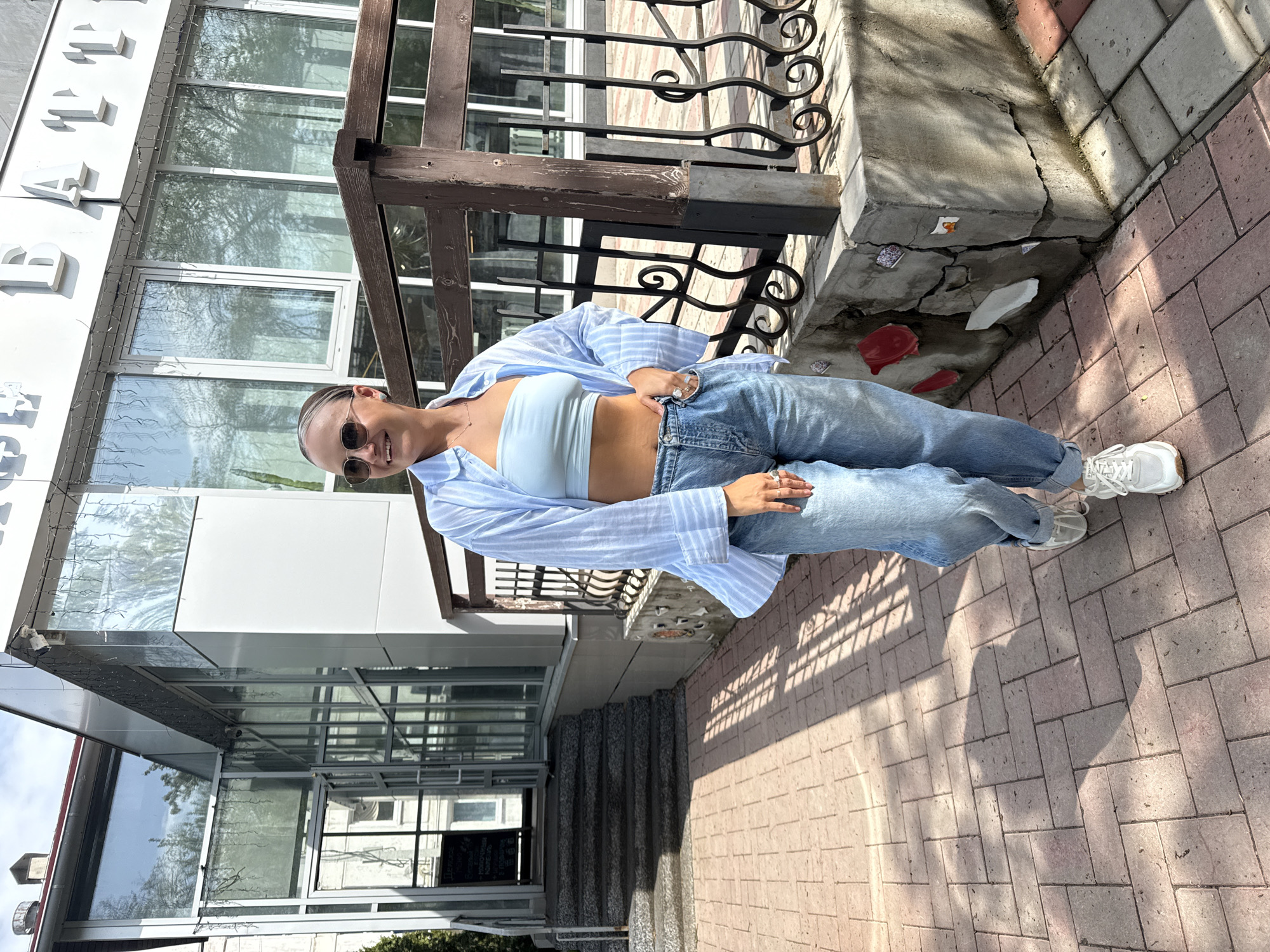 A woman is standing on a brick sidewalk in Chaikovsky, Russia. She is wearing sunglasses, a light blue top, light blue jeans, and a light blue and white striped shirt. She has her hands on her hips. Behind her is a building with glass windows and a dark brown railing. There is also an ornate black metal railing. The sidewalk is made of pink and gray bricks. The picture is taken during the daytime.