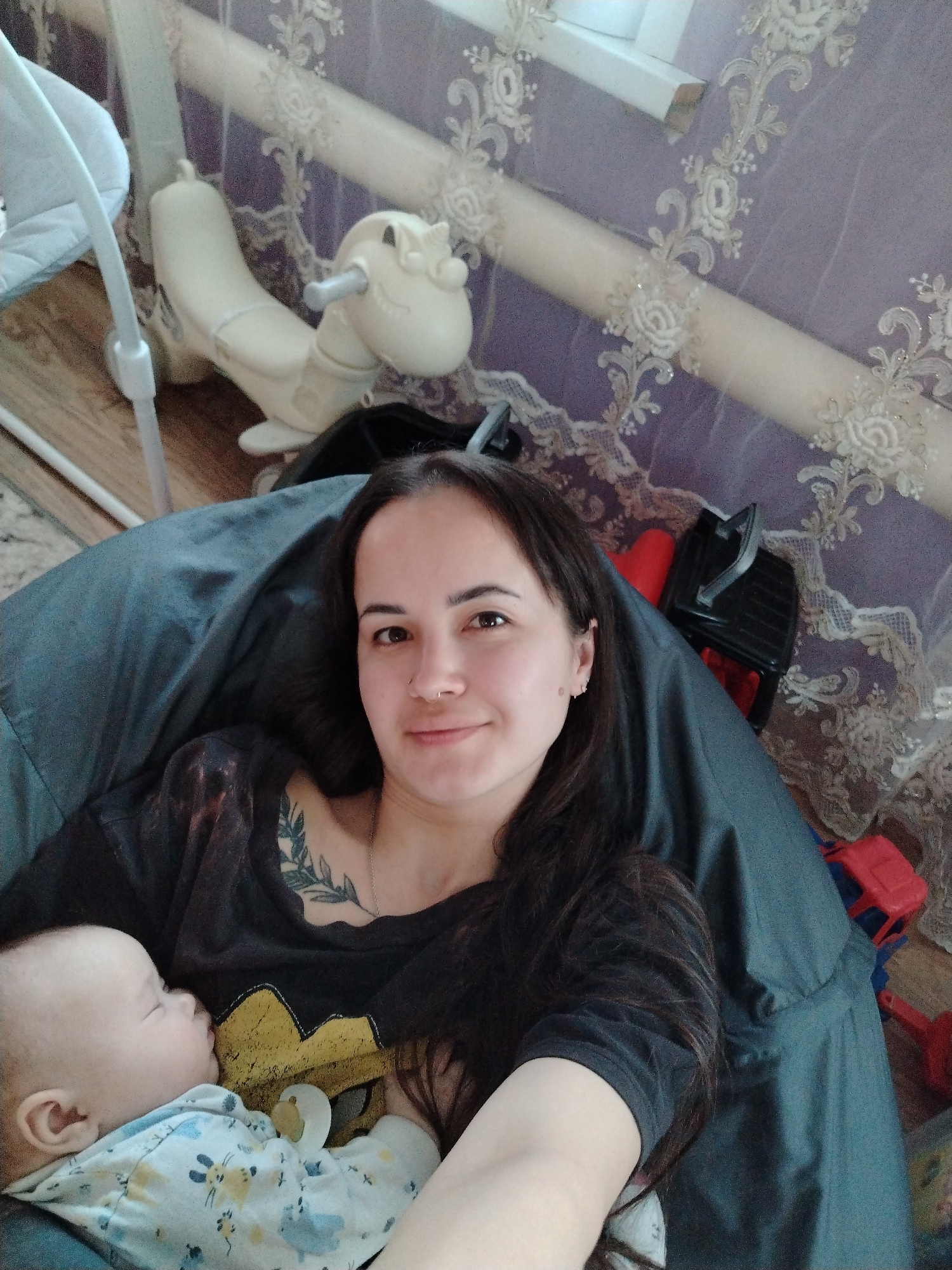 A medium shot shows a woman with long dark hair, a nose ring, and a tattoo on her chest, smiling at the camera. She is sitting in a large, dark gray beanbag chair, holding a sleeping baby dressed in patterned pajamas. Behind her is a window with lace curtains, a white rocking horse toy, a baby swing, and some children's toys. The location is Гожан, Russia.