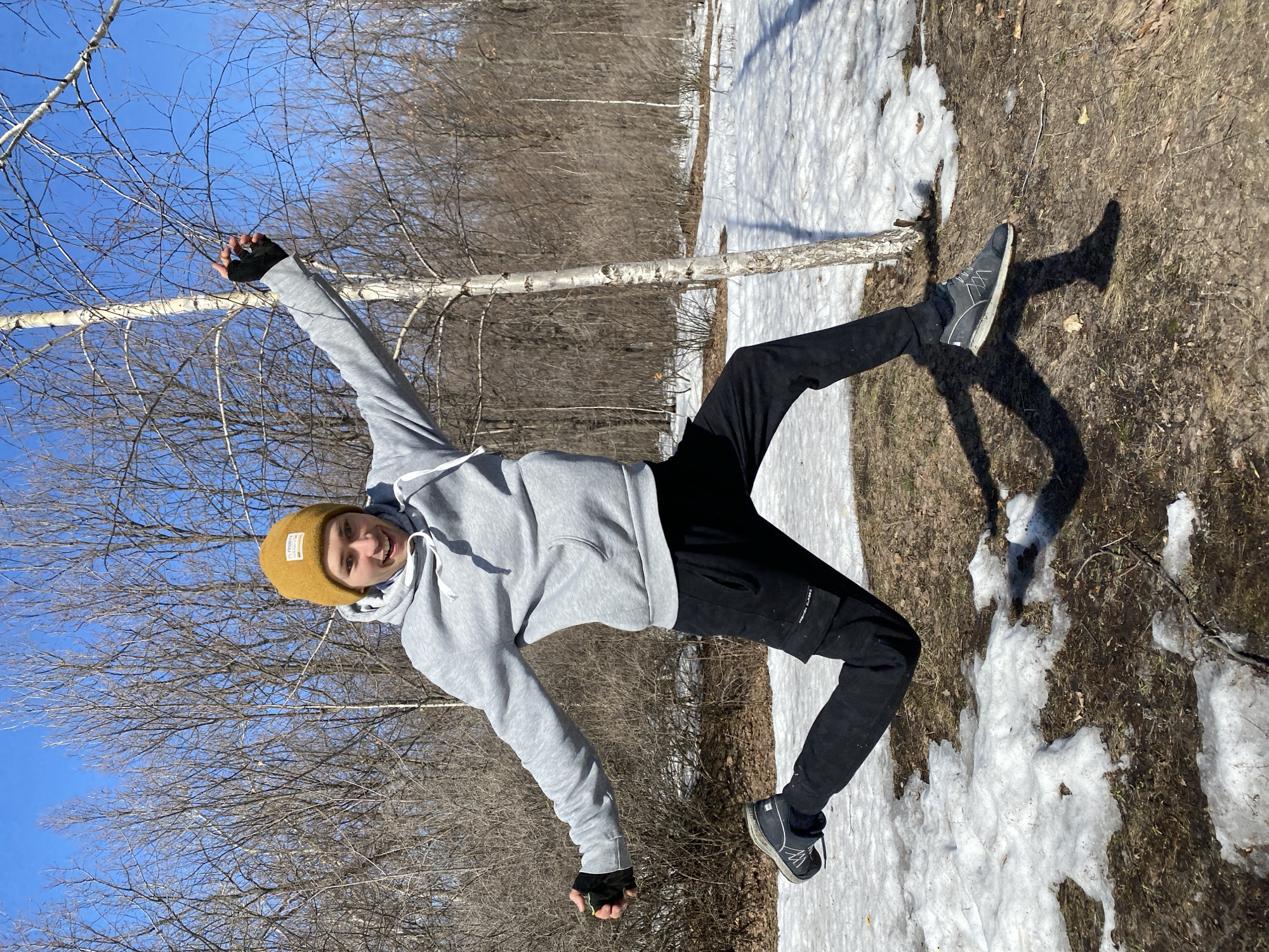 A young man is photographed outdoors, with his body positioned diagonally across the frame, appearing to be captured in a dynamic pose. He wears a light grey hooded sweatshirt, black pants, fingerless gloves, a yellow beanie, and grey athletic shoes. The background shows a landscape with bare trees and patches of snow, likely indicating late winter or early spring. The location is not identifiable as Ulyanovsk, Russia based on the image.