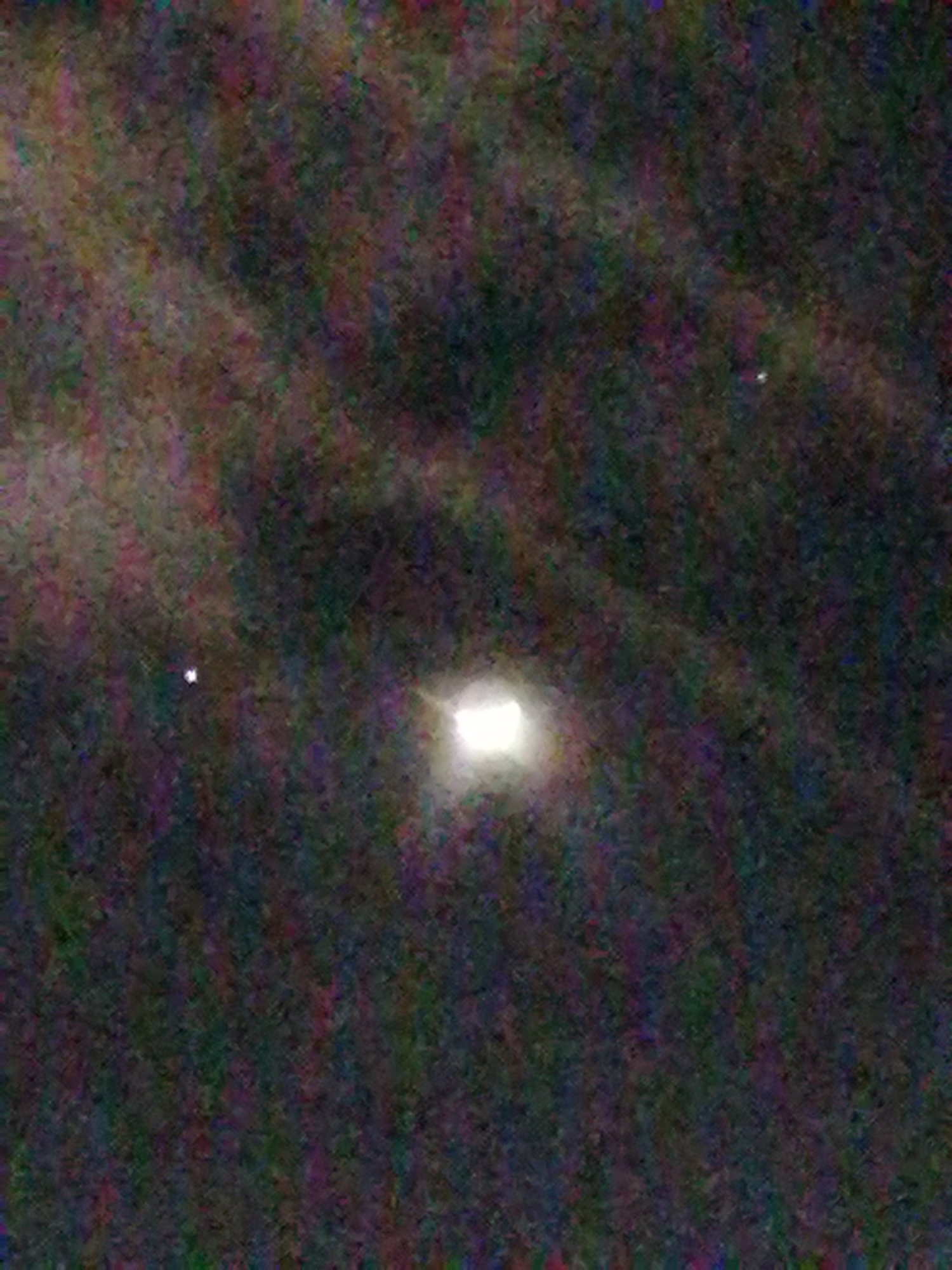 The image appears to show a poorly lit night scene, potentially in a somewhat rural or less brightly illuminated part of Da Lat, Vietnam. In the center of the image is a bright, square-shaped light source, possibly a streetlamp or a building light. The background is dark and blurry, suggesting it is the night sky with a hint of cloud cover or vegetation. Two smaller points of light are scattered in the periphery, possibly stars. The image is noisy and lacks clarity.
