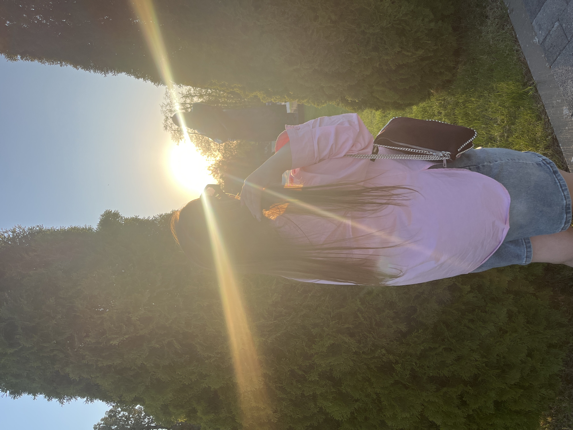 The image depicts a person standing outdoors in what appears to be a park or garden. The location may be in or near Novopokrovskaya, Russia. The person is wearing a light pink shirt and denim shorts. They have long, dark hair. The sun is visible in the background, creating a strong backlight and glare. A dark-colored bag with a chain strap is slung over the person's shoulder. The surrounding environment includes green bushes or trees and a paved area. The focus is primarily on the person and their immediate surroundings.